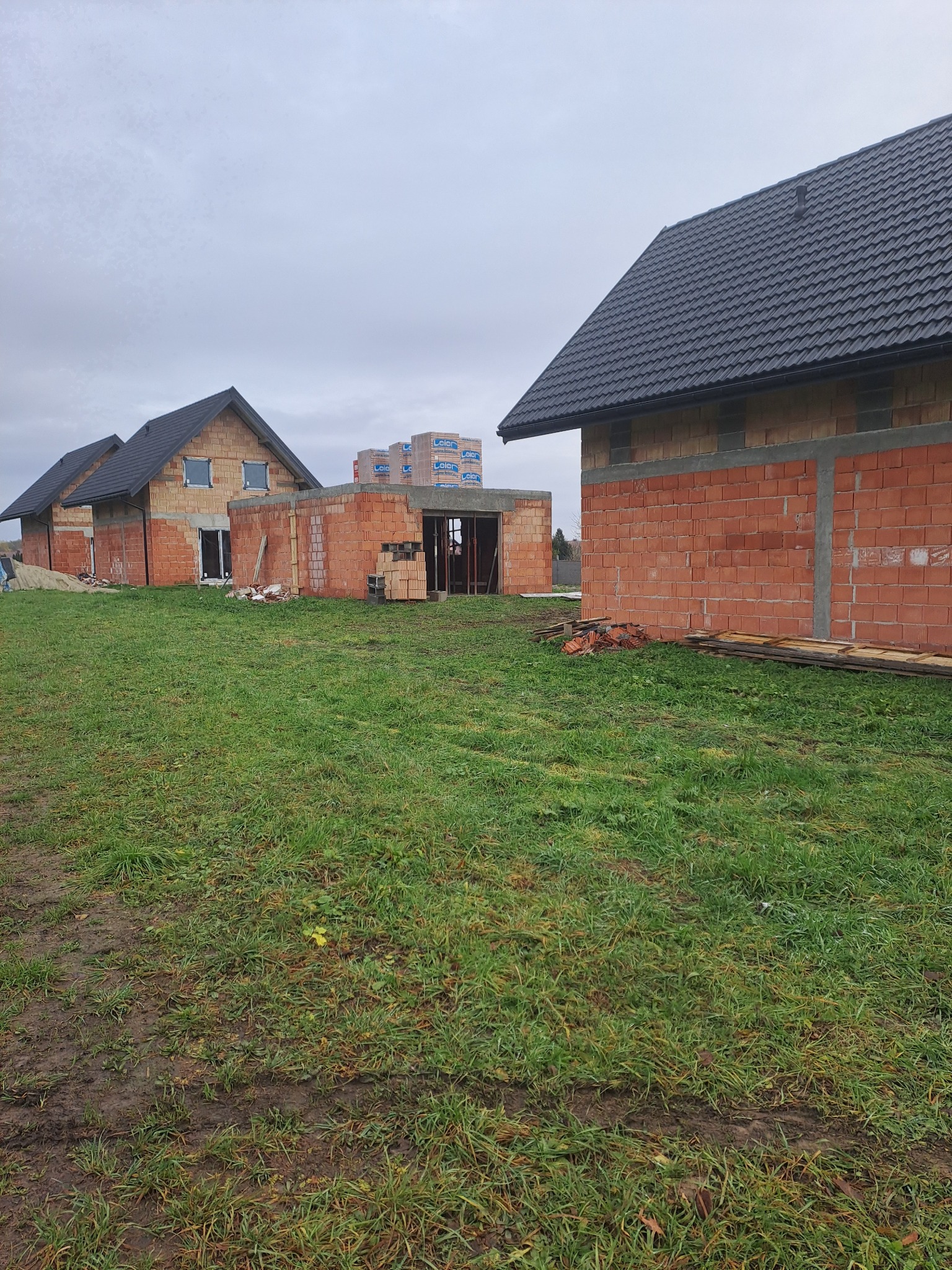 Budowa domów jednorodzinnych w stanie surowym otwartym z czerwonej cegły. Widoczne ściany, dachy z czarnej dachówki i palety z materiałami budowlanymi.