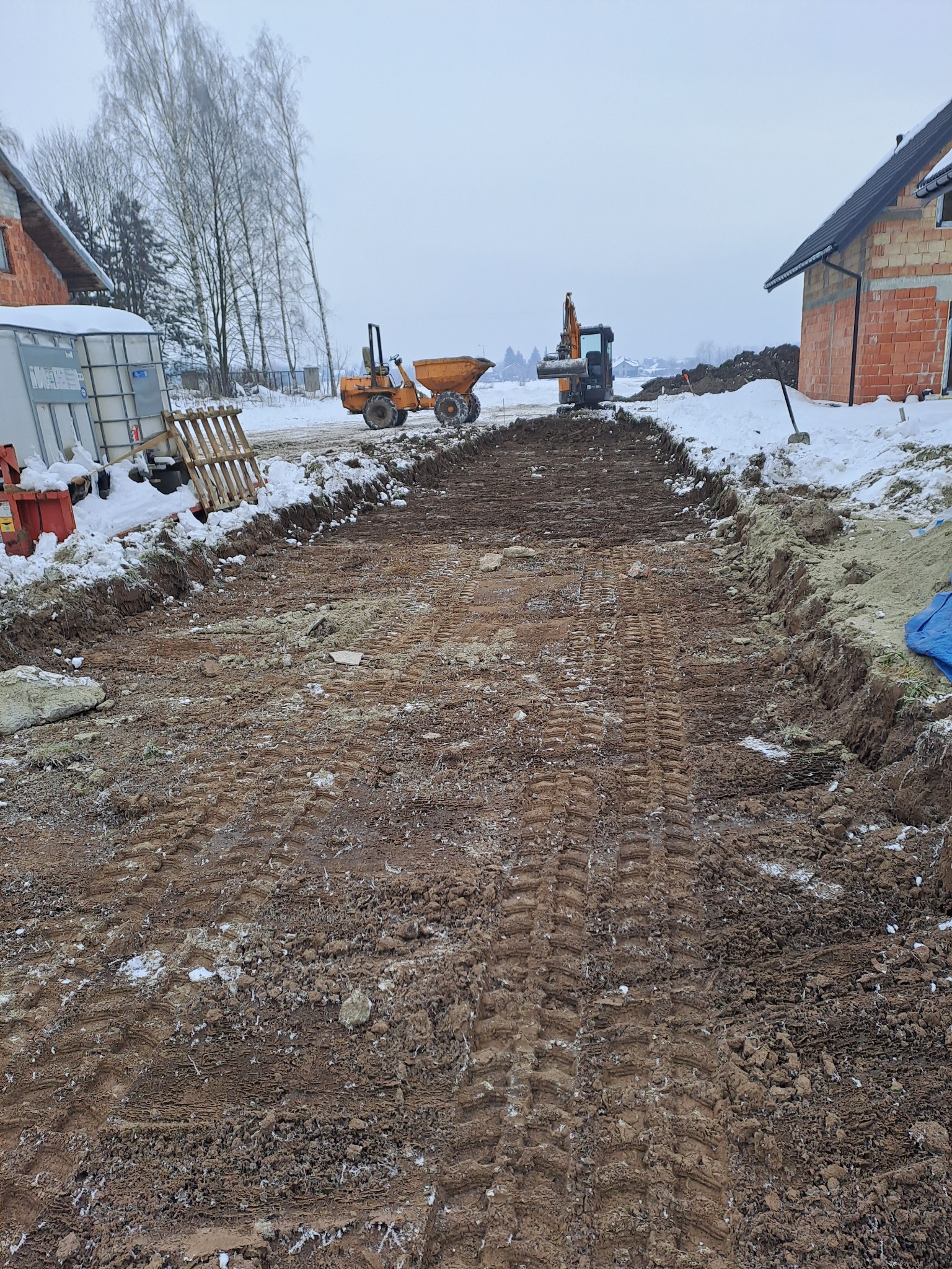 Wykop pod fundamenty domu w zimowej scenerii. Widoczny wykopany pas ziemi, maszyny budowlane i fragmenty murowanych ścian w tle. Ślady opon na ziemi.