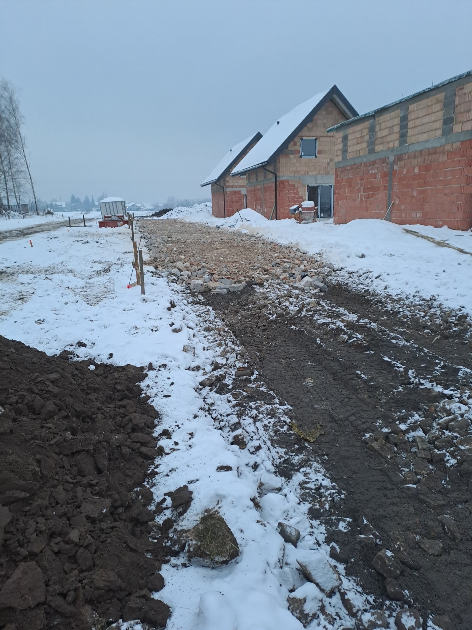 Zimowy krajobraz budowy domu z czerwonej cegły. Widoczne fundamenty, gruz i ślady opon na śniegu. Pochmurne niebo.