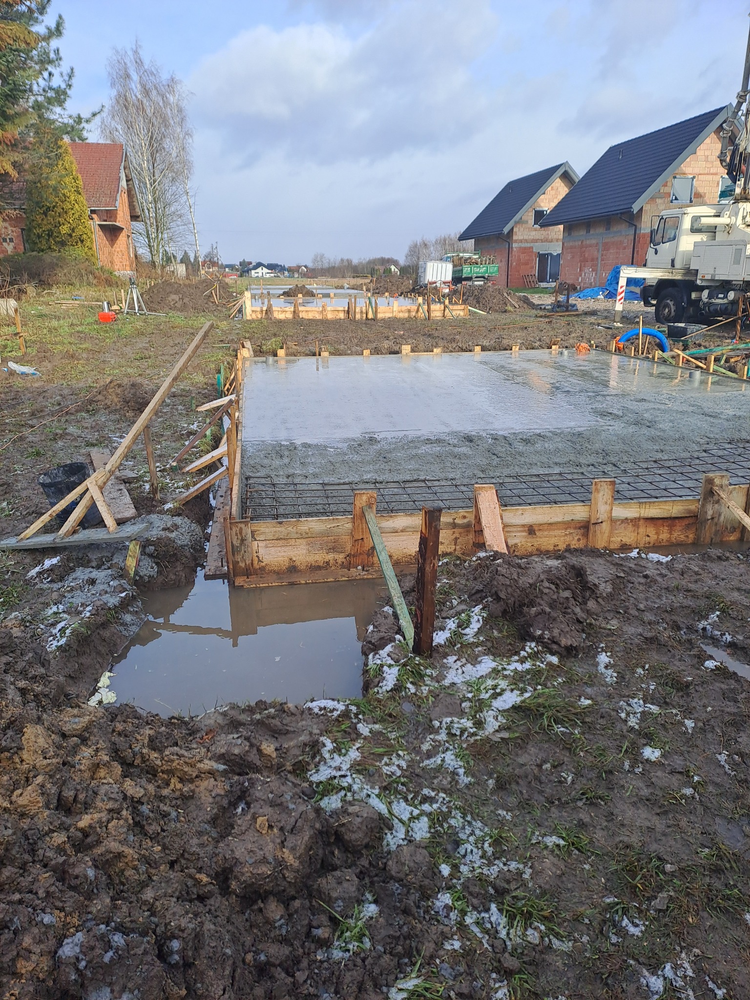 Zalany betonem fundament zbrojony siatką, w drewnianym szalunku. W tle domy w trakcie budowy i maszyna budowlana. Błotniste podłoże z resztkami śniegu.
