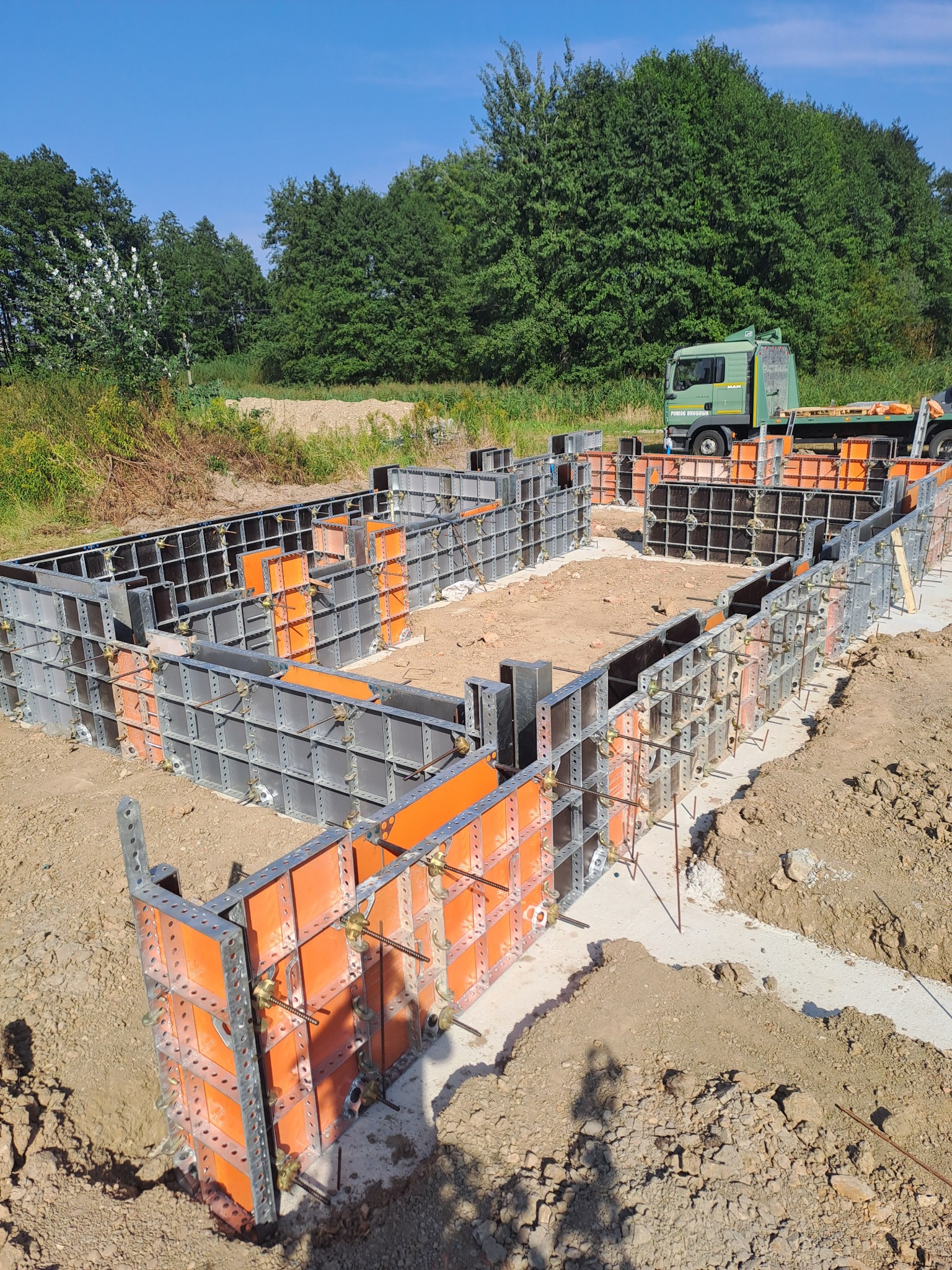 Szare szalunki z pomarańczowymi elementami, tworzące formę fundamentów domu na ziemi, z zielonym samochodem ciężarowym w tle i drzewami w słoneczny dzień.