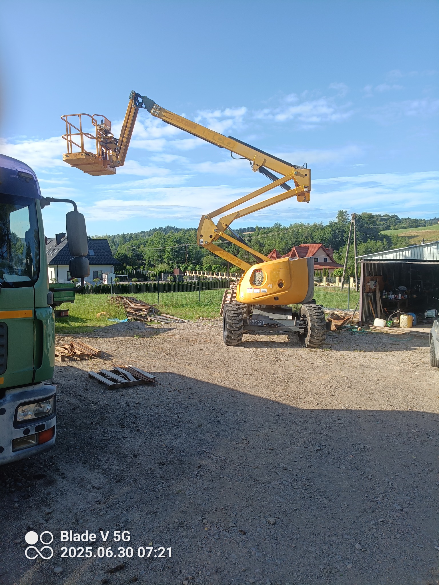 Żółty podnośnik koszowy na nierównym terenie, obok zaparkowany zielony samochód ciężarowy. W tle domy i zalesione wzgórza w słoneczny dzień.