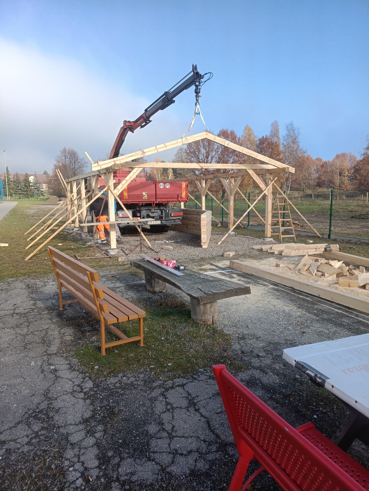 Montaż drewnianej konstrukcji z użyciem dźwigu samochodowego. Widoczny pracownik w pomarańczowym ubraniu. W tle drzewa i niebieskie niebo.