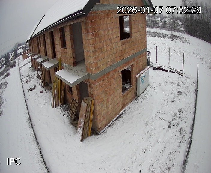 Wznoszenie szeregowca z czerwonej cegły w zimowej scenerii. Widoczne rusztowania i deski. Ujęcie z kamery przemysłowej z datą i godziną w prawym górnym rogu.