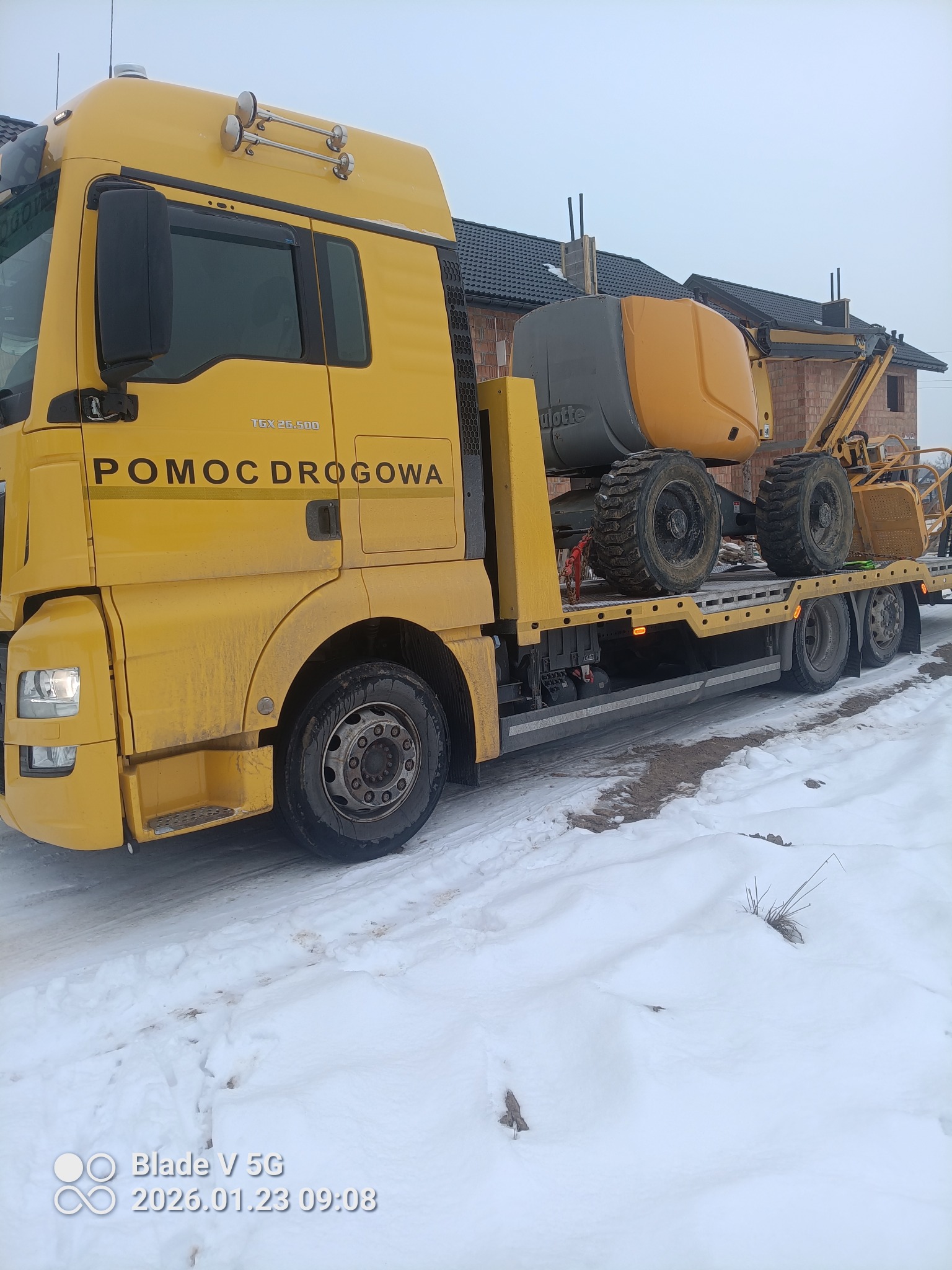 Żółta laweta z podnośnikiem koszowym na tle budowy domu w Jastrzębi. Transport maszyn budowlanych. Usługi budowlane i wynajem sprzętu.