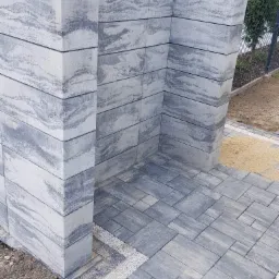 Wykończony narożnik z szarej kostki brukowej o wzorze imitującym kamień, obrzeże z białego grysu, widoczny fragment trawnika i ogrodzenia.