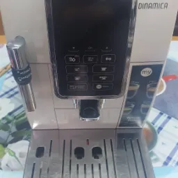 Ekspres do kawy DeLonghi Dinamica w kolorze kremowym, z panelem sterowania z ikonami napojów, widoczny element spieniacza do mleka i tacka ociekowa z metalowymi kratkami, na tle stołu z obrusem...