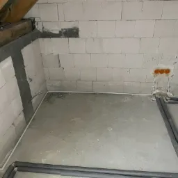 Surowy stan poddasza w trakcie prac: widoczne przewody elektryczne w białej osłonie biegnące po ścianie z bloczków, puszki elektryczne w pomarańczowej obudowie osadzone w ścianie oraz rury grzewcze...