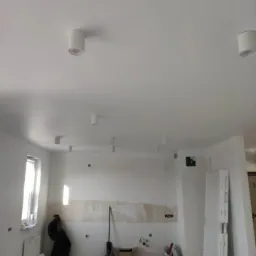 Wnętrze w trakcie remontu z zamontowanymi cylindrycznymi lampami sufitowymi w kolorze białym, widoczne ślady po pracach malarskich na ścianie, okno z białymi ramami i grzejnikiem pod nim.