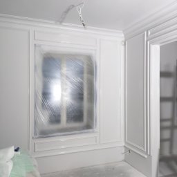 UWAGA!!Wykonanie gładzi,malowanie!i td... - Wnętrze po świeżym malowaniu na biało, okno zabezpieczone folią, widoczne listwy dekoracyjne, pistolet do malowania leży na podłodze.