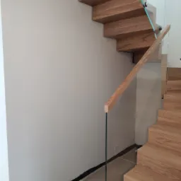 Nowoczesne schody drewniane z szklaną balustradą w trakcie montażu w mieszkaniu w Warszawie, widoczne zabezpieczenia podłogi.