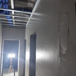 UWAGA!!Wykonanie gładzi,malowanie!i td... - Ściana w trakcie nakładania gładzi, widoczne fragmenty nierówności obok wygładzonej powierzchni, metalowa konstrukcja sufitu z niebieskimi rurami instalacji, lampa budowlana na trójnogu w tle.