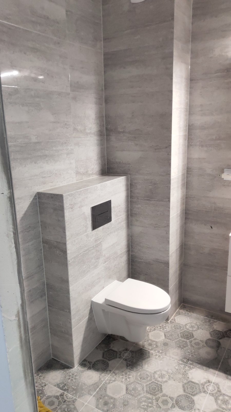 Nowoczesna toaleta podwieszana z zabudową stelaża, wykończona szarą płytką imitującą kamień, z ozdobną podłogą w heksagonalne wzory.