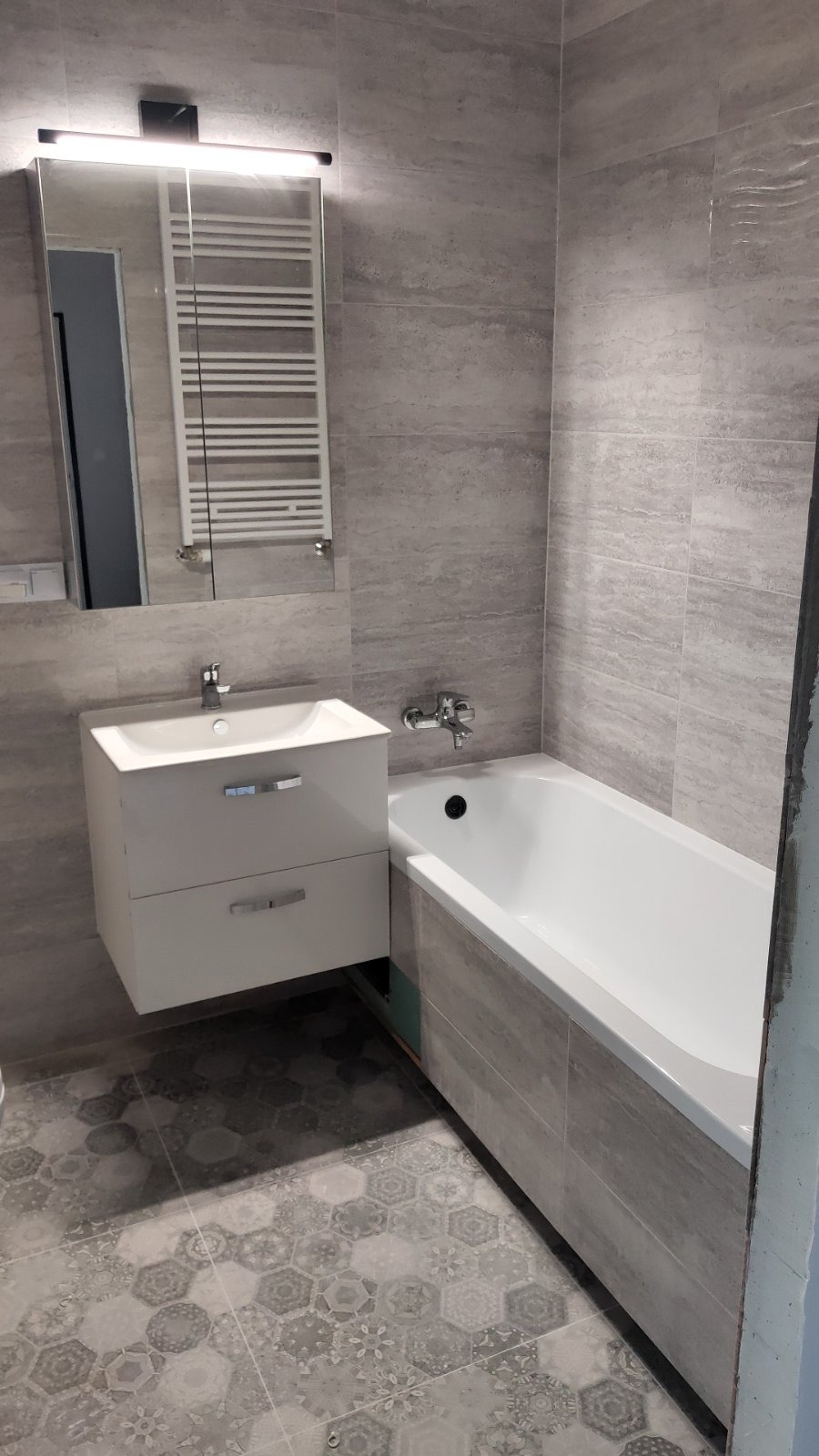 Nowoczesna łazienka w szarościach z płytkami imitującymi kamień na ścianach, białą wanną i umywalką z szafką, wzorzysta podłoga.