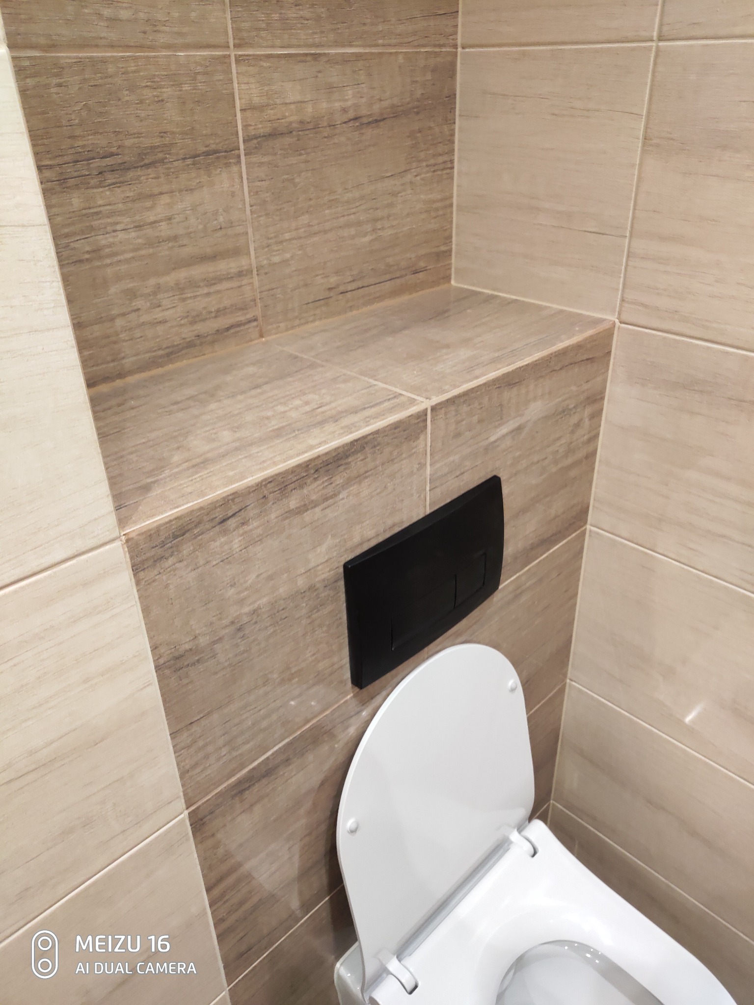 Wykończona toaleta z drewnopodobnymi płytkami ceramicznymi na ścianach i zabudowie, widoczny biały sedes z otwartą klapą i czarny panel spłuczki.