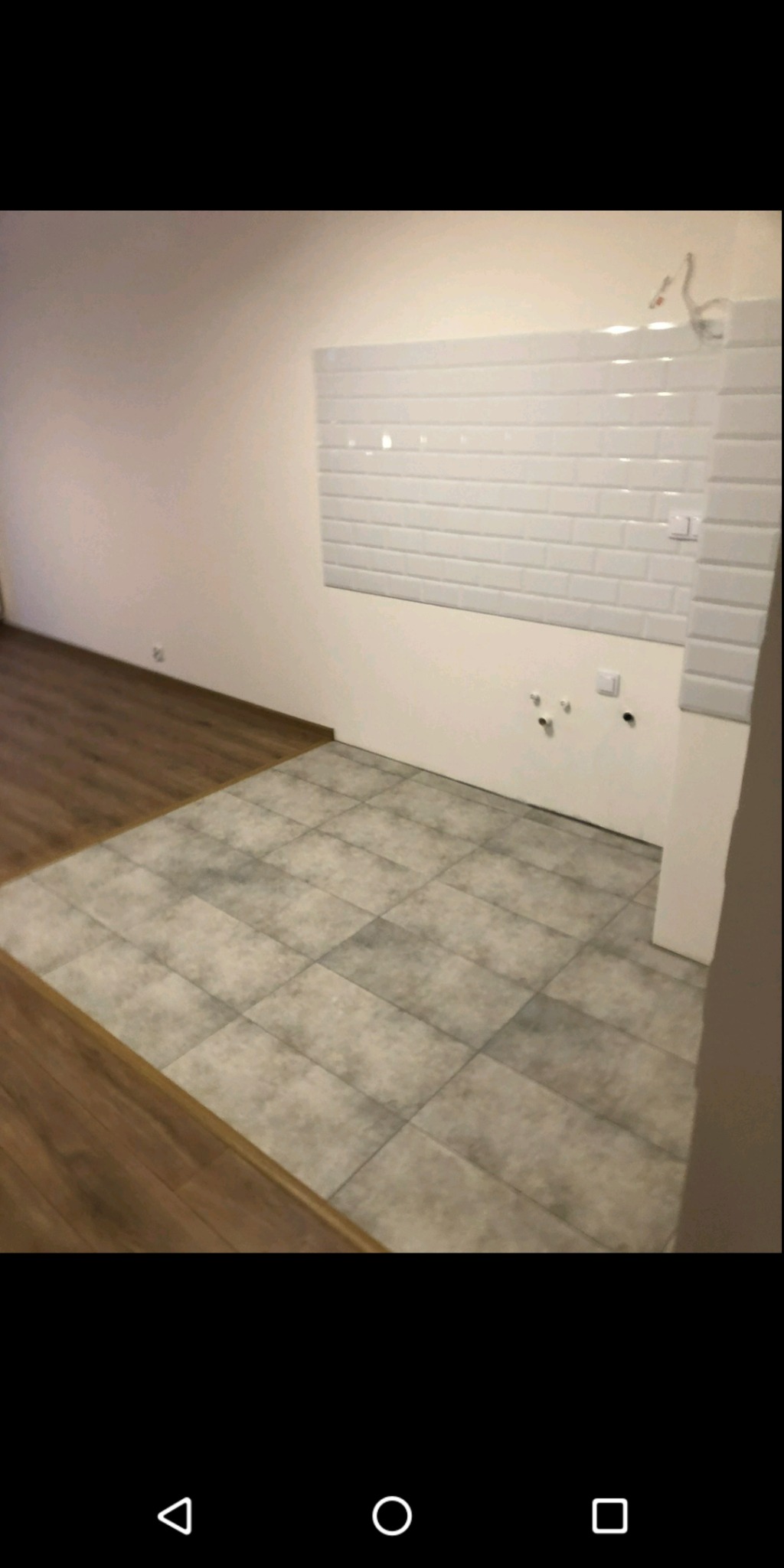 Fragment wnętrza po remoncie: szare płytki podłogowe przechodzą w drewniany parkiet, białe ściany z białymi kaflami w stylu cegiełek, widoczne wyprowadzenia instalacji elektrycznej i wodnej.