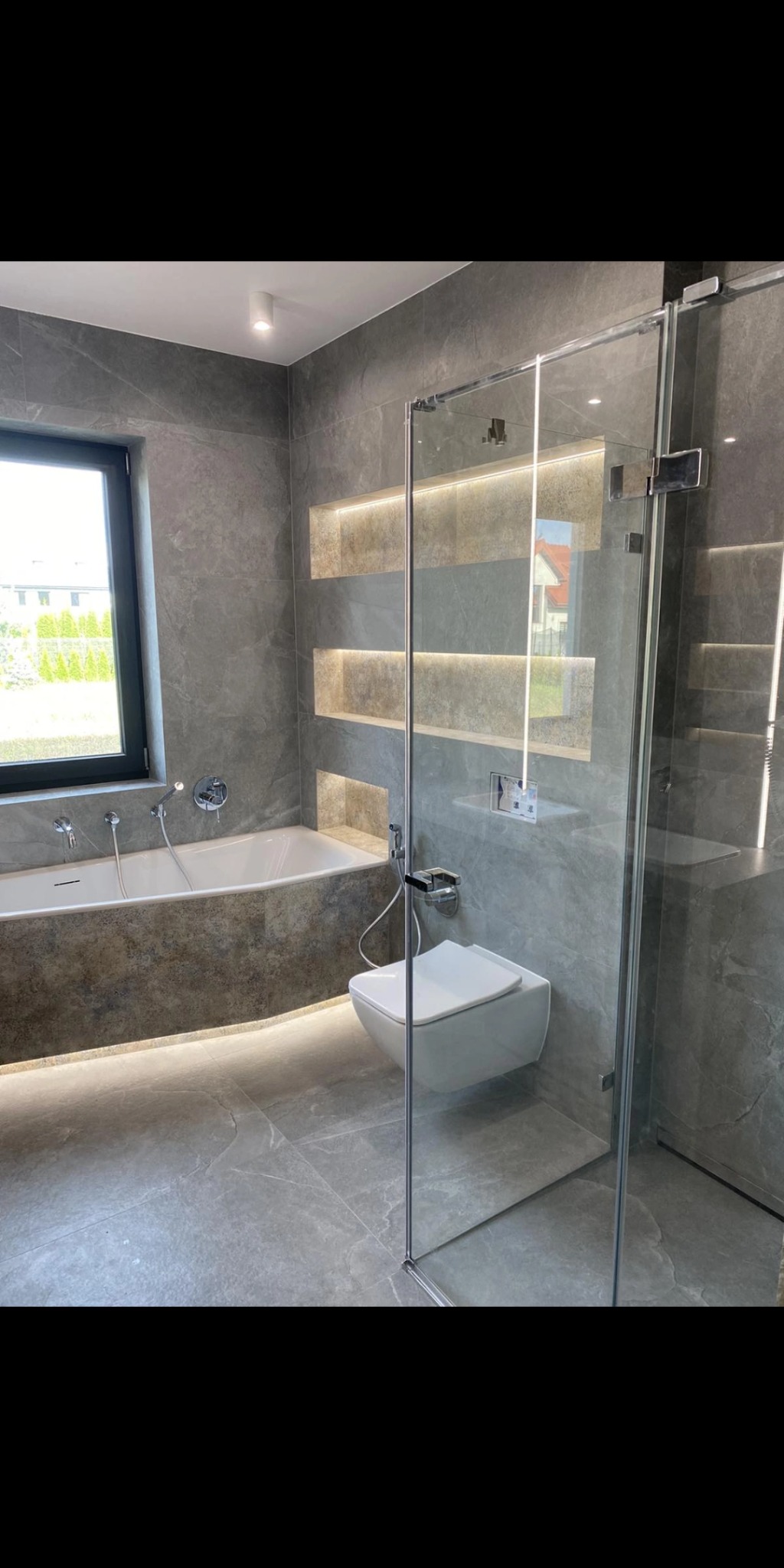 Nowoczesna łazienka z szarymi płytkami imitującymi marmur, podświetlanymi niszami w ścianie, białą wiszącą toaletą i kabiną prysznicową ze szkła hartowanego.