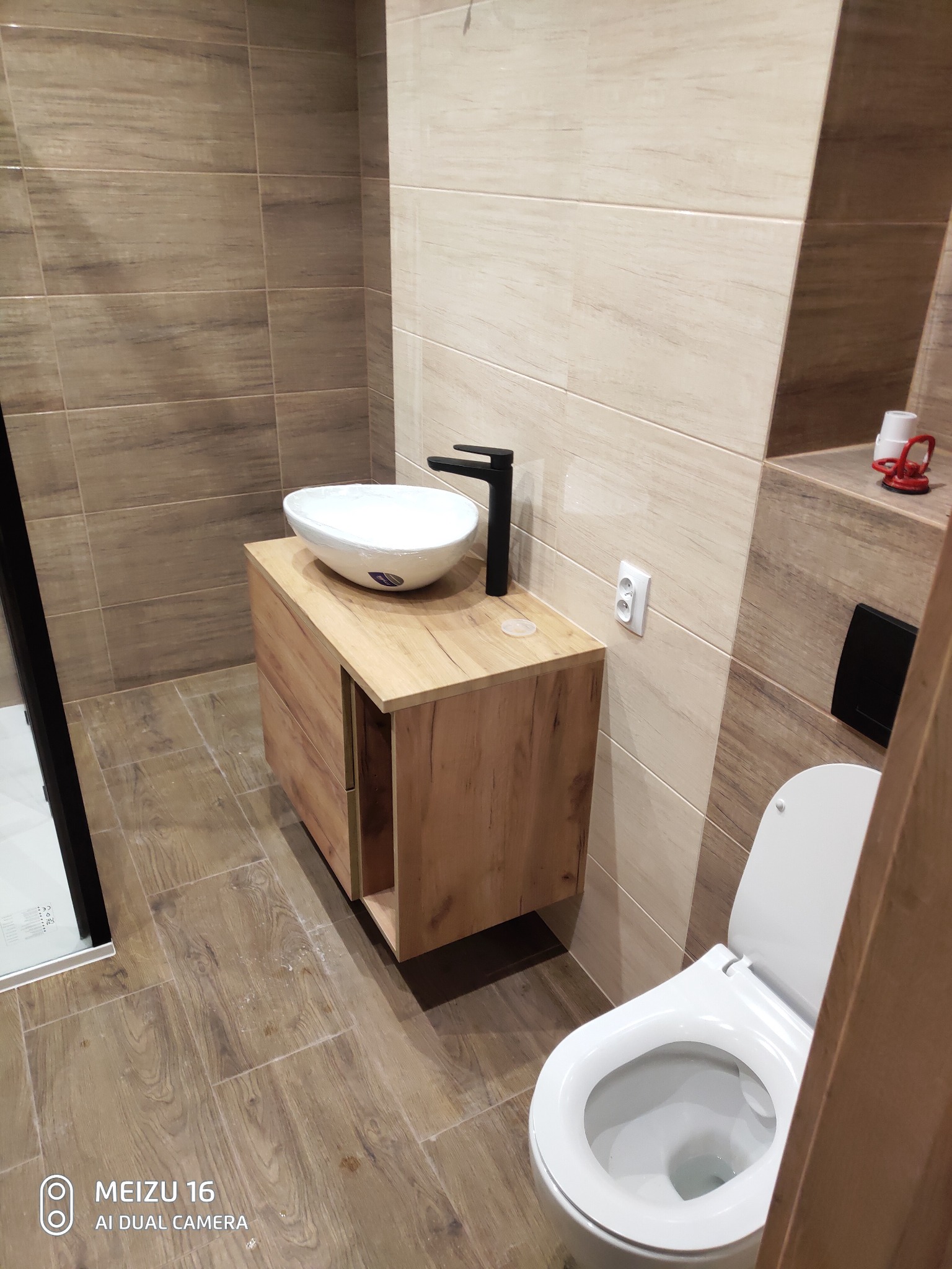 Nowoczesna łazienka z umywalką nablatową w folii ochronnej na szafce z drewnopodobnym fornirem, czarną armaturą i toaletą w kolorze białym, wykończona płytkami imitującymi drewno.