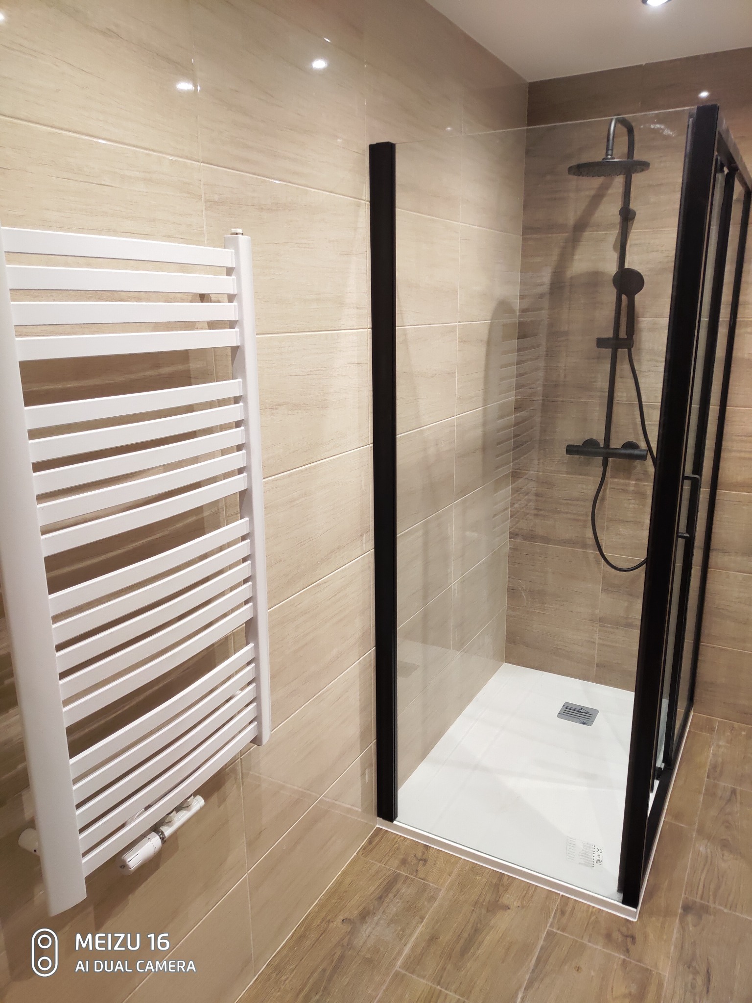 Nowoczesna kabina prysznicowa z czarnymi profilami, biały grzejnik drabinkowy na ścianie z beżowych płytek imitujących drewno.