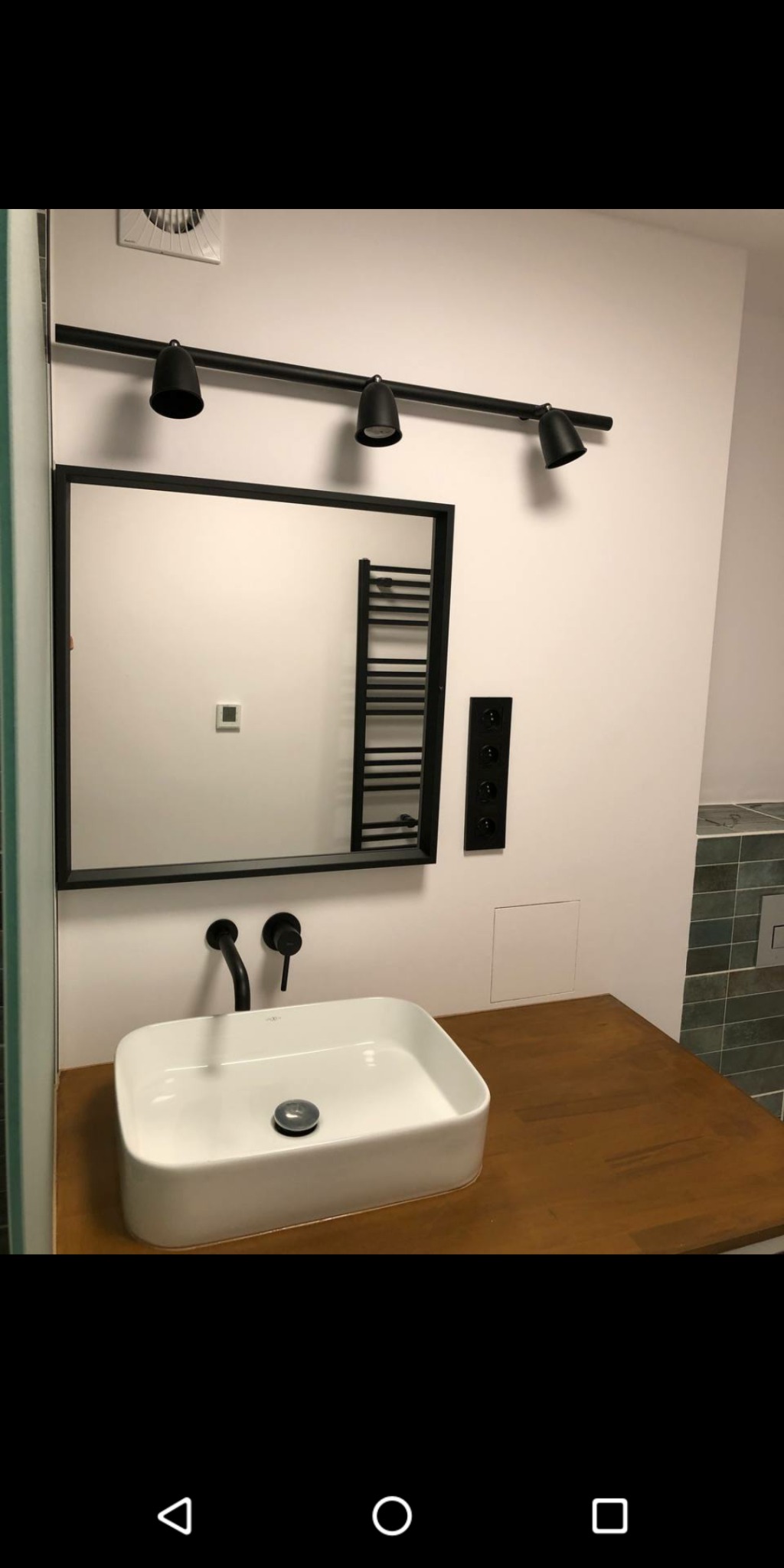 Nowoczesna łazienka z białą, prostokątną umywalką nablatową, czarną armaturą, lustrem w czarnej ramie, oświetleniem szynowym z trzema reflektorami i grzejnikiem drabinkowym w tle. Blat drewniany...