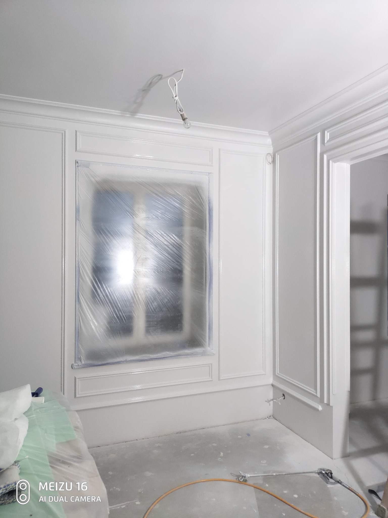 Wnętrze po świeżym malowaniu na biało, okno zabezpieczone folią, widoczne listwy dekoracyjne, pistolet do malowania leży na podłodze.