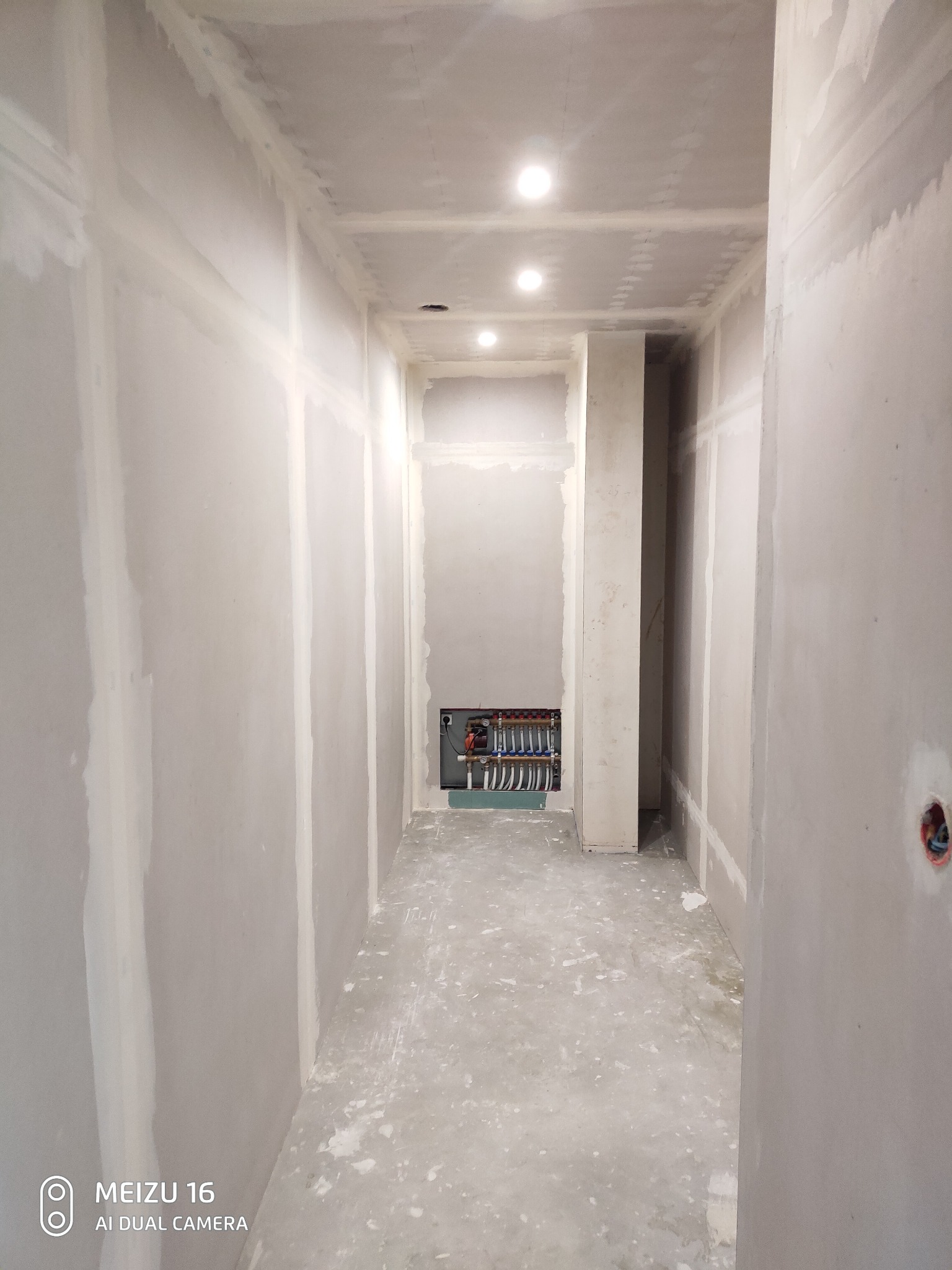 Korytarz w trakcie prac wykończeniowych w Warszawie: ściany przygotowane do malowania, widoczne łączenia płyt gipsowo-kartonowych i instalacja grzewcza w zagłębieniu ściany.