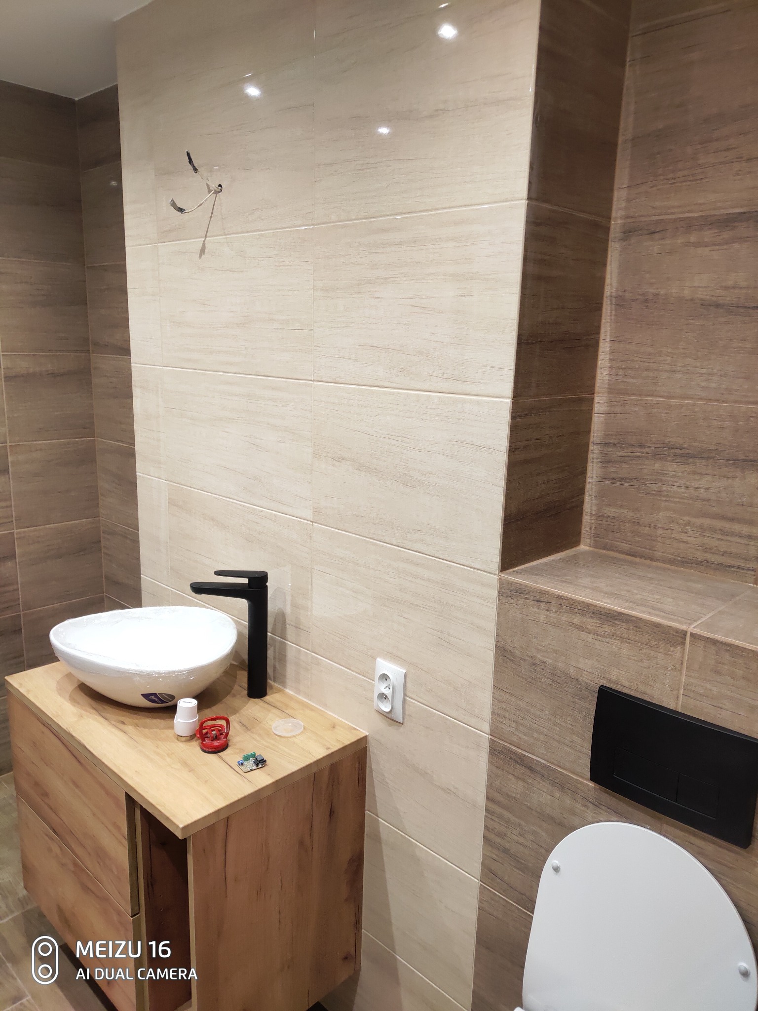 Nowoczesna łazienka w trakcie wykańczania: beżowe i brązowe płytki imitujące drewno, biała umywalka nablatowa z czarną baterią, drewniana szafka pod umywalką, widoczne narzędzia i materiały...