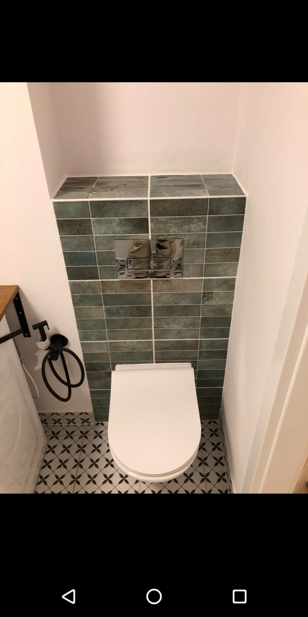 Wykończona toaleta z białym sedesem, zielono-szarą mozaiką na ścianie i czarno-białą wzorzystą podłogą, widoczny fragment pralki i czarny bidet.