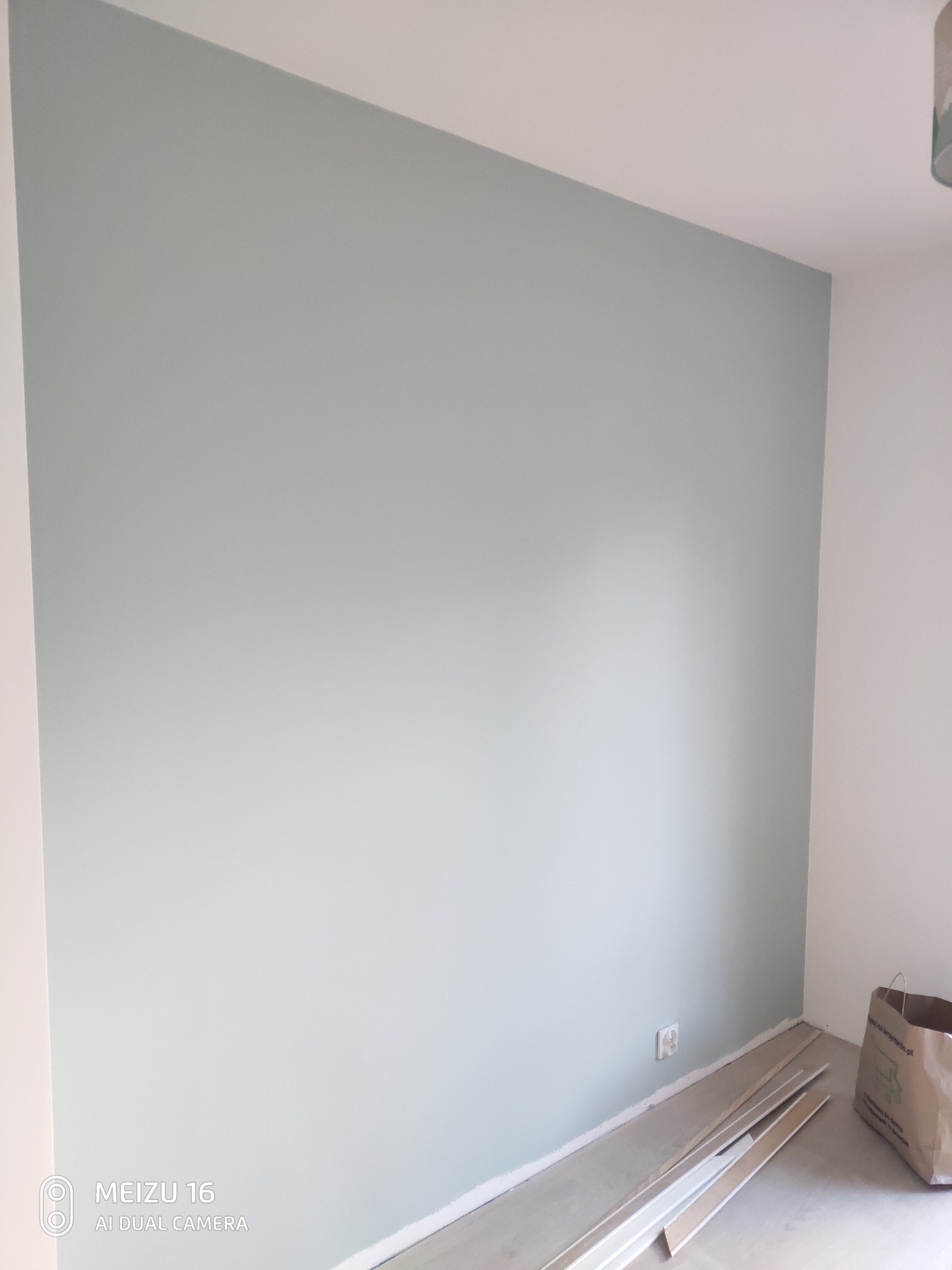 Świeżo pomalowana ściana w odcieniu pastelowej zieleni, widoczne listwy przypodłogowe i torba papierowa w rogu pomieszczenia, gniazdko elektryczne na ścianie.