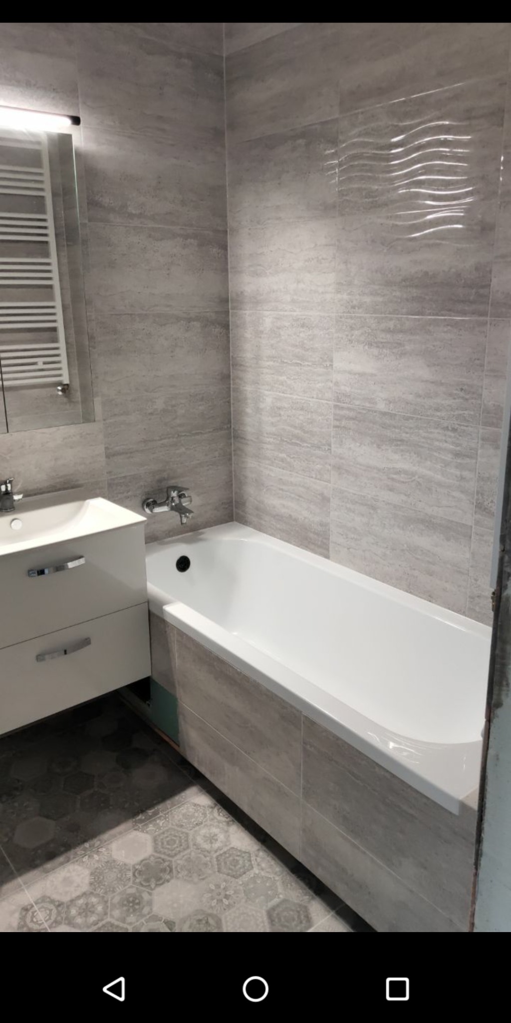 Wykończona łazienka z wanną obudowaną płytkami imitującymi kamień, umywalką z szafką, grzejnikiem drabinkowym i heksagonalnymi płytkami podłogowymi.