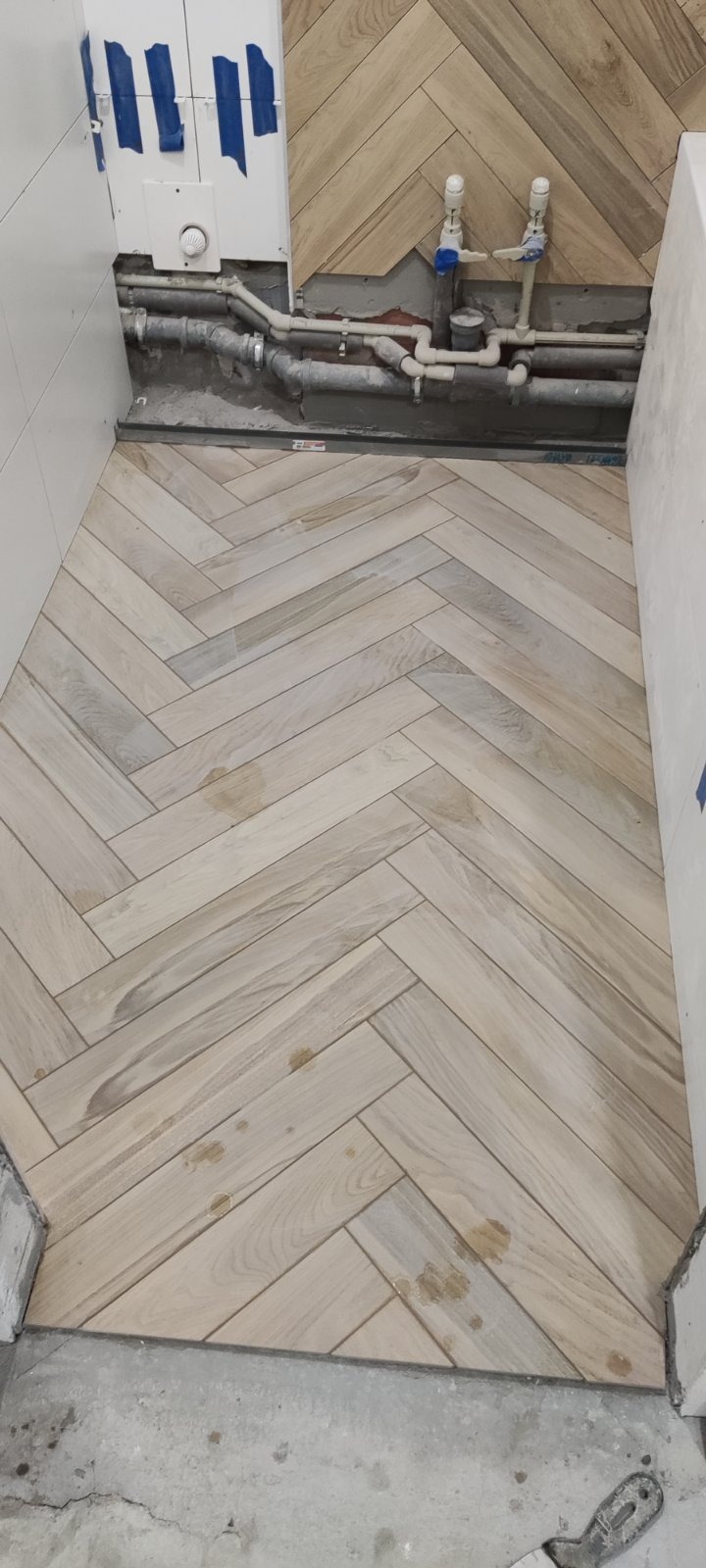 Ułożona pod kątem jodełki jasna podłoga z płytek imitujących drewno w trakcie prac wykończeniowych w łazience, widoczne niedociągnięcia przy ścianach i rurach.