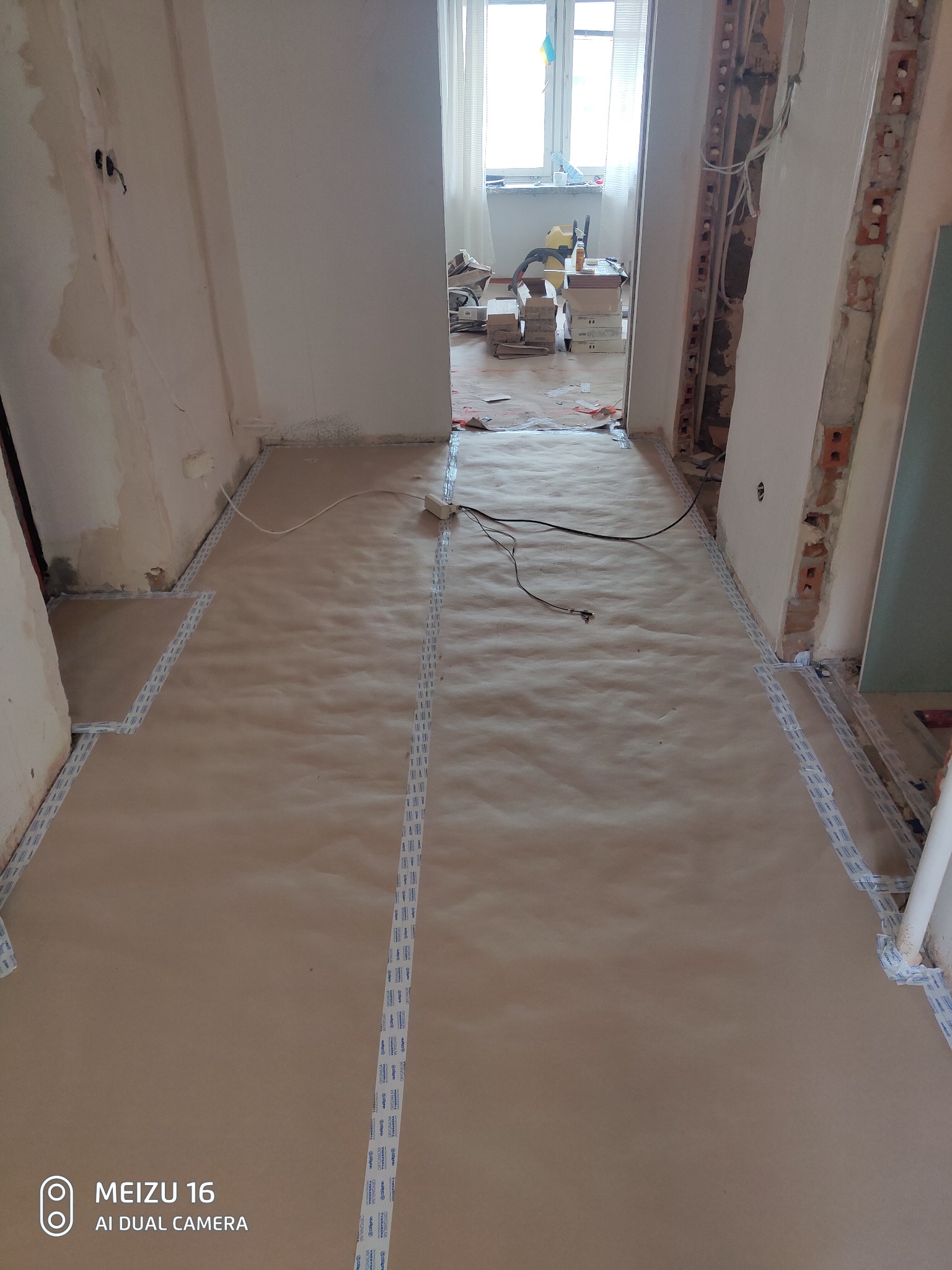 Przygotowanie podłoża w remontowanym mieszkaniu: podłoga zabezpieczona brązowym papierem i taśmą ochronną, widoczne ślady po instalacjach elektrycznych w ścianie, okno w tle.