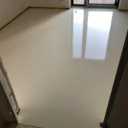 Jasna, błyszcząca podłoga żywiczna w nowym pomieszczeniu, zabezpieczona taśmą malarską przy ścianach i oknach, z widocznym fragmentem surowej podłogi przy progu.