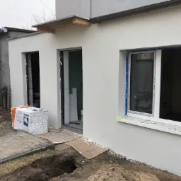 Wstępnie ocieplona ściana budynku z otworami okiennymi i drzwiowymi, widoczne bloczki styropianowe na palecie, przygotowane do montażu okna i drzwi, świeżo nałożony tynk.