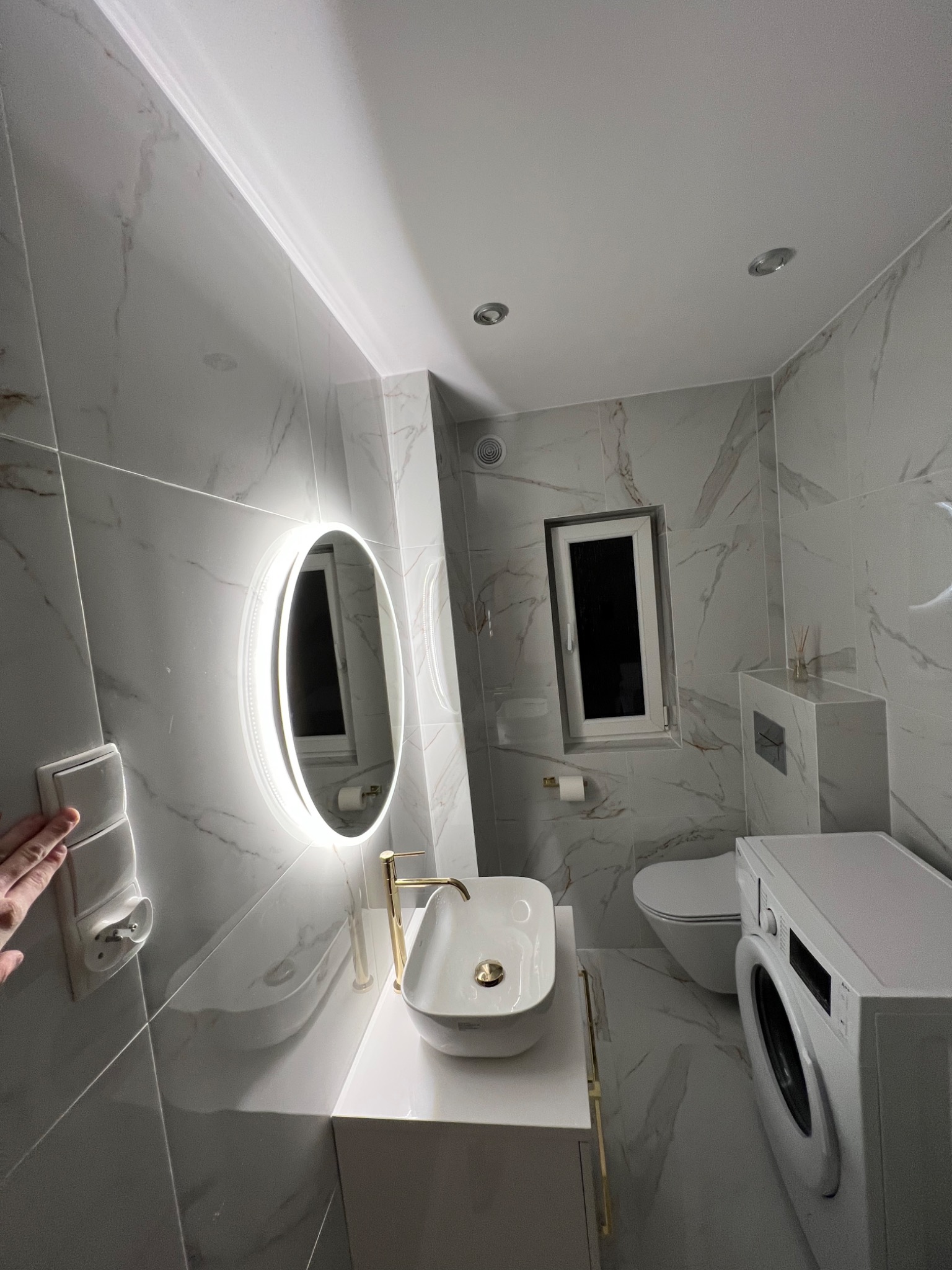 Nowoczesna łazienka wykończona w bieli i szarości z marmurkowym wzorem na płytkach, owalne lustro z podświetleniem LED, biała umywalka nablatowa ze złotą armaturą, toaleta i pralka.