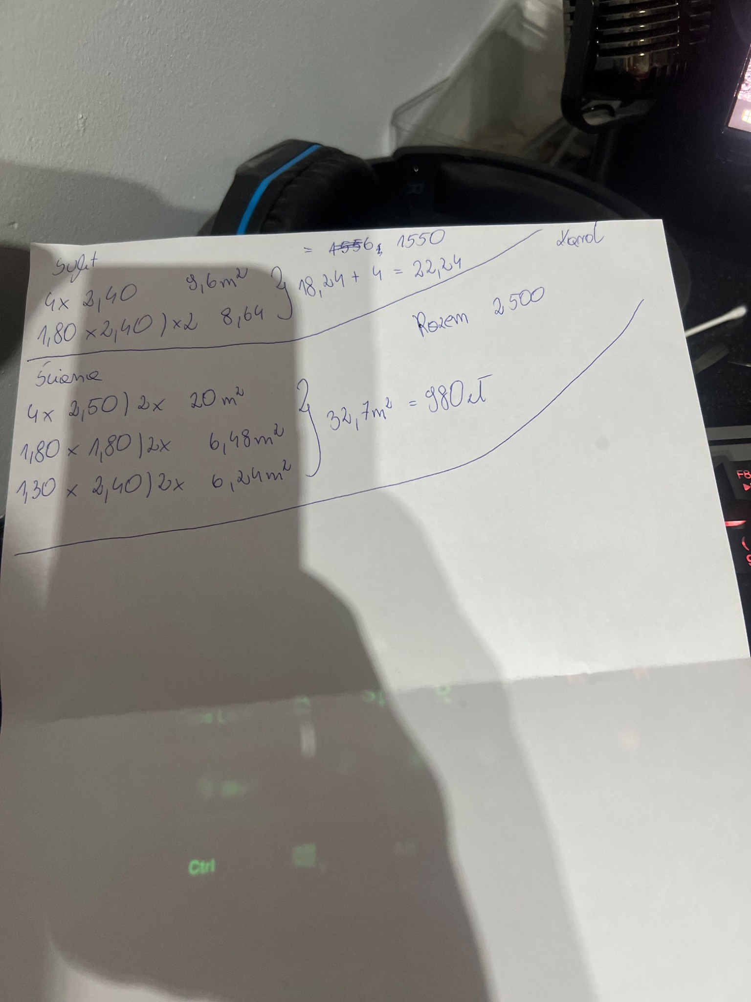 Notatki z obliczeniami powierzchni sufitu i ścian, prawdopodobnie w kontekście remontu lub wykończenia, zapisane na kartce papieru leżącej na klawiaturze komputera.