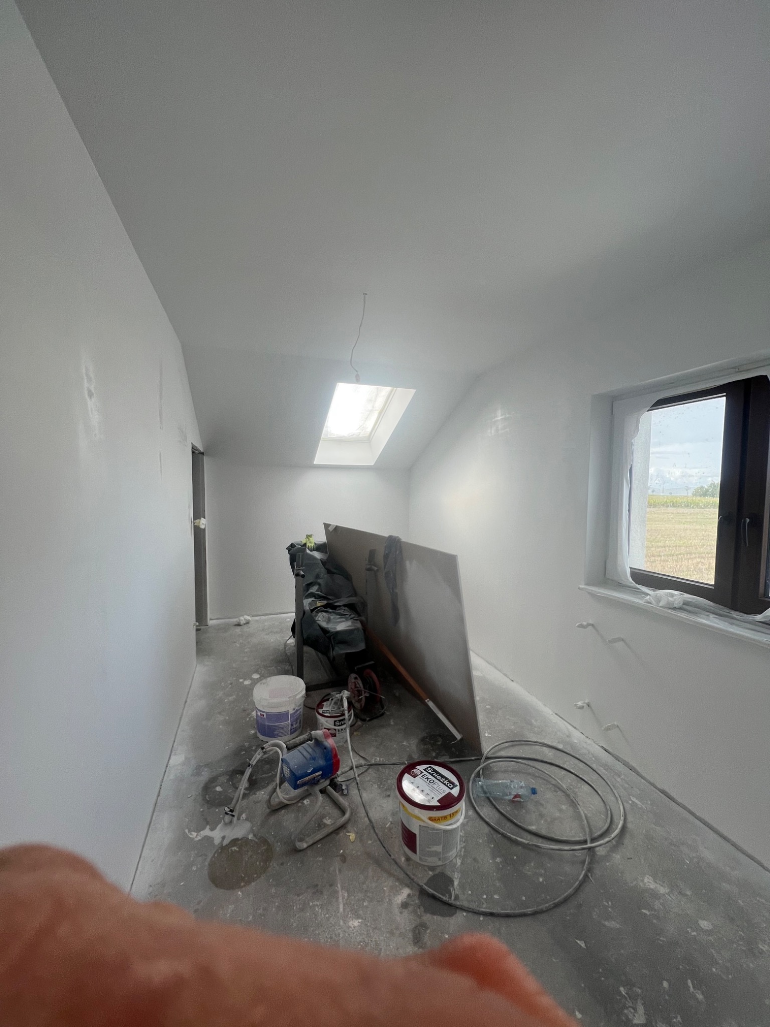Pokój na poddaszu w trakcie malowania natryskowego, widoczny agregat malarski, puszki z farbą Śnieżka EKO Plus i zabezpieczone okno dachowe.