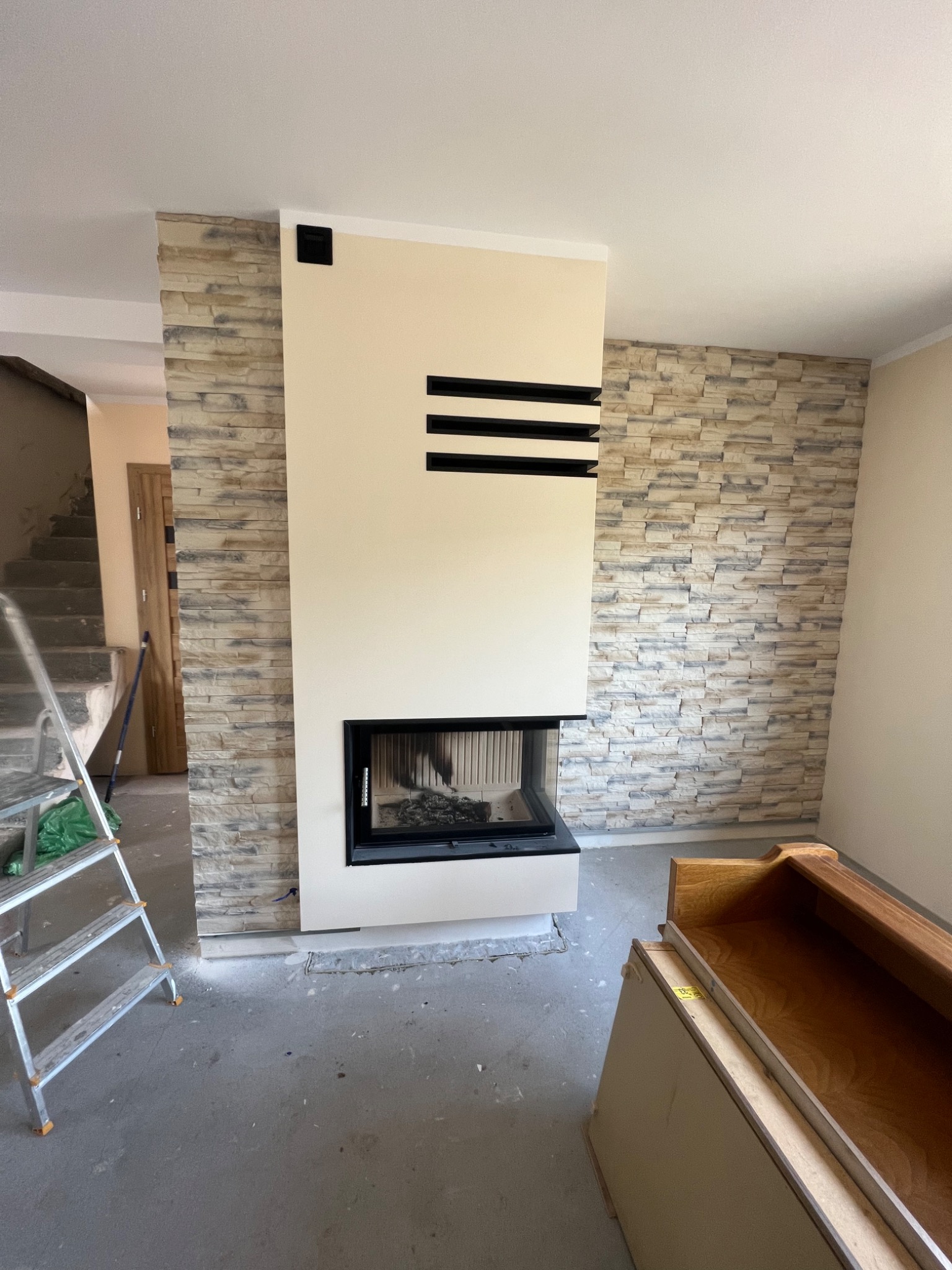 Wnętrze w trakcie wykańczania: kominek z obudową z beżowej płyty z poziomymi czarnymi wstawkami, ściana wyłożona dekoracyjnym kamieniem, widoczna drabina malarska i elementy mebli przed montażem.