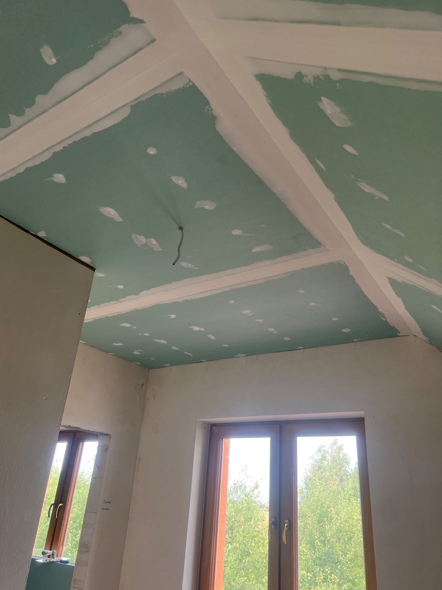 Poddasze w trakcie remontu, widoczne płyty gipsowo-kartonowe na suficie ze szpachlowanymi łączeniami, dwa okna z widokiem na drzewa.