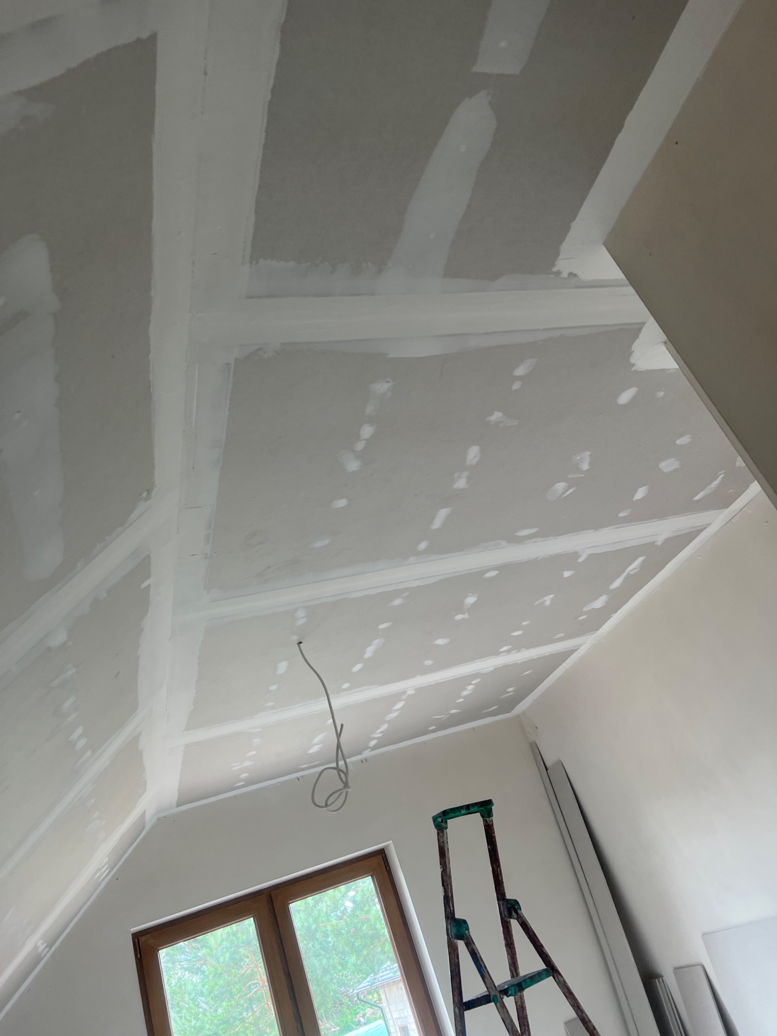 Sufit podwieszany z płyt gipsowo-kartonowych na poddaszu, widoczne łączenia szpachlowane, drabina malarska w rogu pomieszczenia, okno dachowe z widokiem na zieleń.