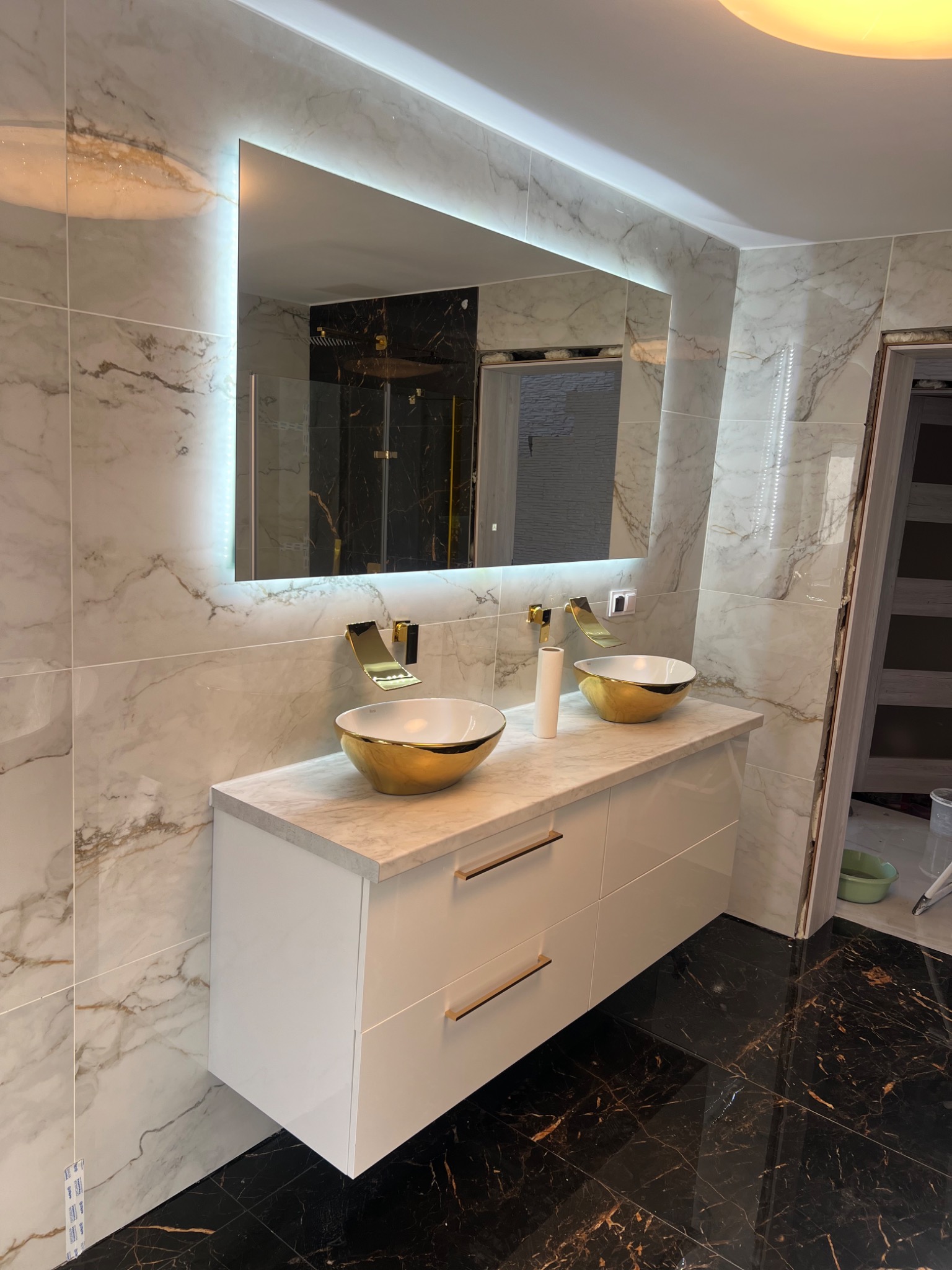 Nowoczesna łazienka z marmurowymi płytkami ściennymi, podwójną umywalką w złotych misach i podświetlanym lustrem, czarna podłoga z żyłkami.