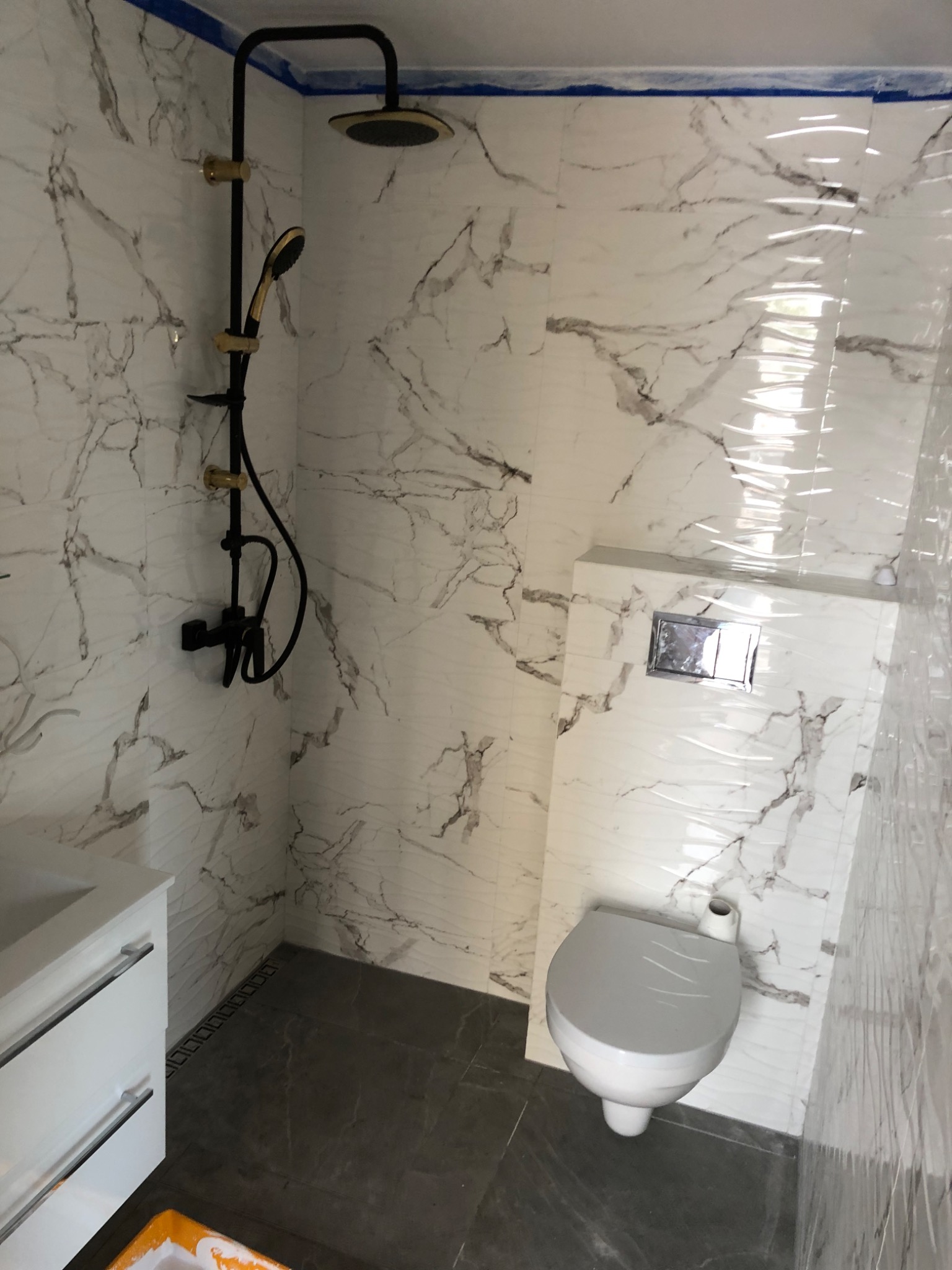 Nowoczesna łazienka w trakcie wykańczania: białe marmurowe płytki, czarna armatura prysznicowa z elementami w kolorze złota, podwieszana toaleta i biała szafka pod umywalkę, zabezpieczone folią...