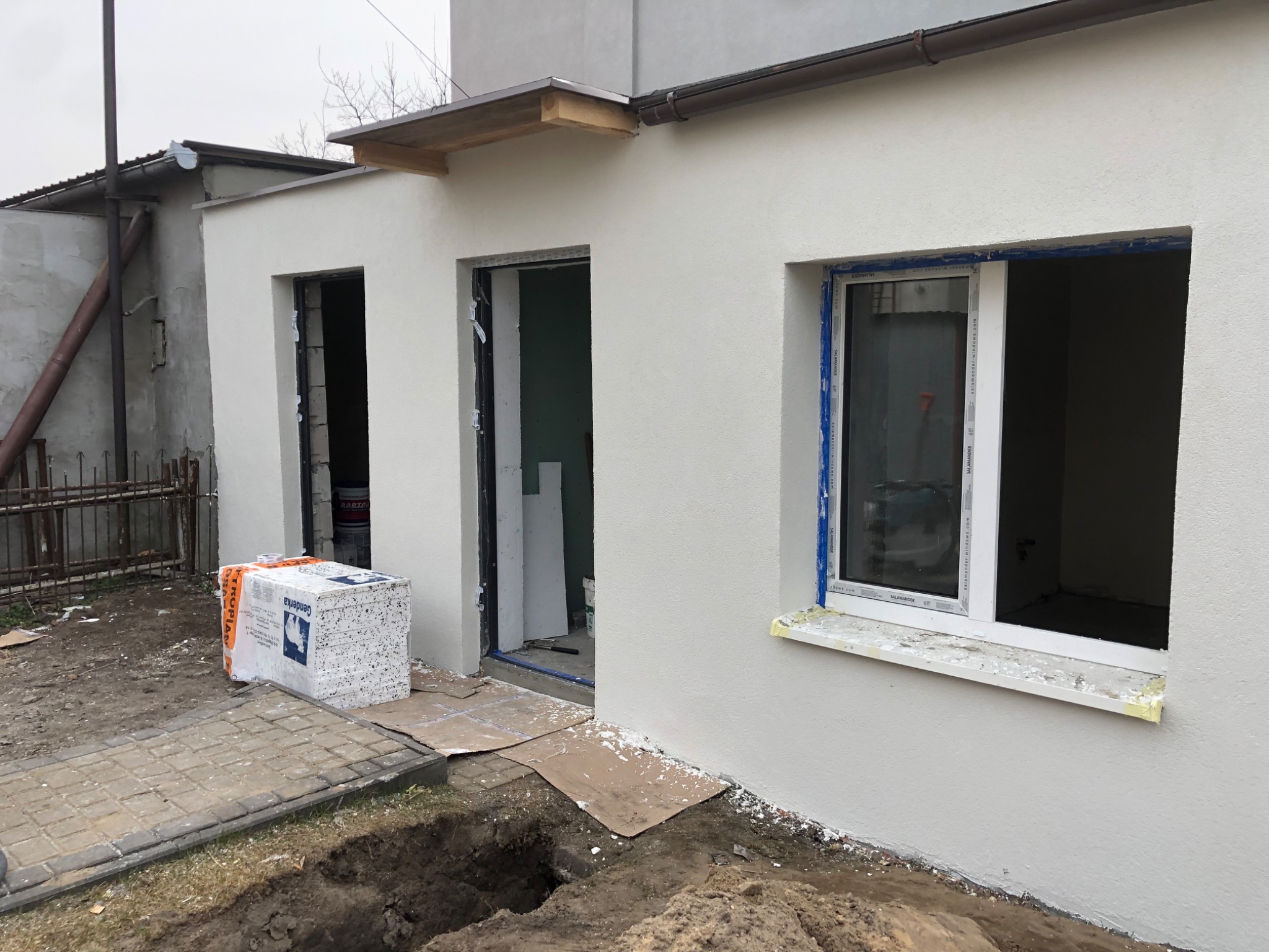 Wstępnie ocieplona ściana budynku z otworami okiennymi i drzwiowymi, widoczne bloczki styropianowe na palecie, przygotowane do montażu okna i drzwi, świeżo nałożony tynk.