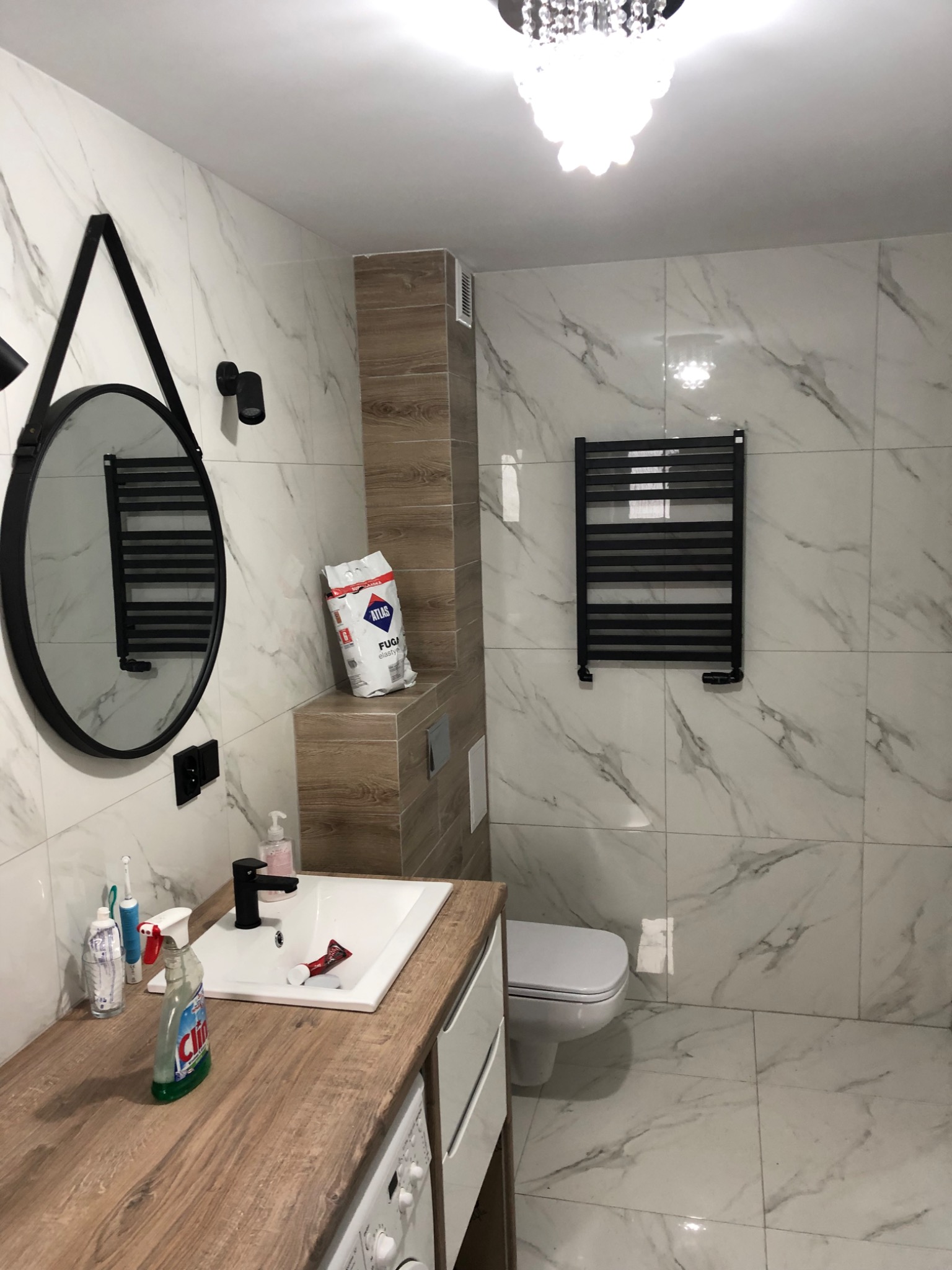 Nowoczesna łazienka z marmurowymi płytkami, okrągłym lustrem na pasku, grzejnikiem drabinkowym i drewnianym blatem z kosmetykami, widoczny worek z fugą Atlas.