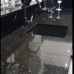 Błyszczący, czarny blat kuchenny z ciemnym zlewem i czarną baterią, uzupełniony o dekoracje i rośliny.