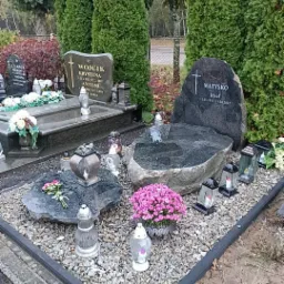 Dwa nagrobki z ciemnego kamienia, otoczone drobnymi kamykami, z lampionami i kwiatami, w tle wysokie tuje.