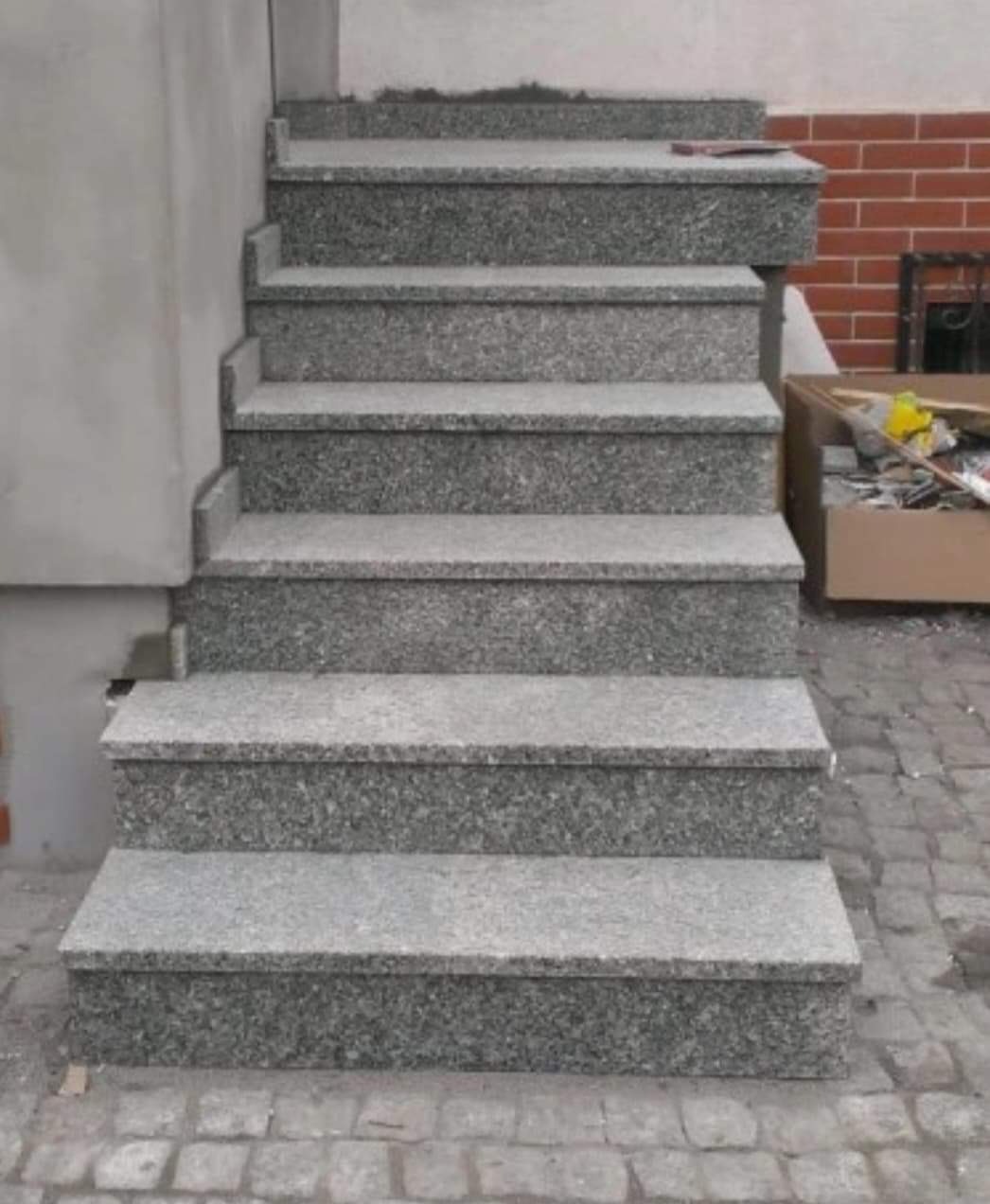 Zewnętrzne schody kamienne z szarego granitu prowadzące do budynku, widok z dołu, w tle fragment muru z cegły i karton z narzędziami.