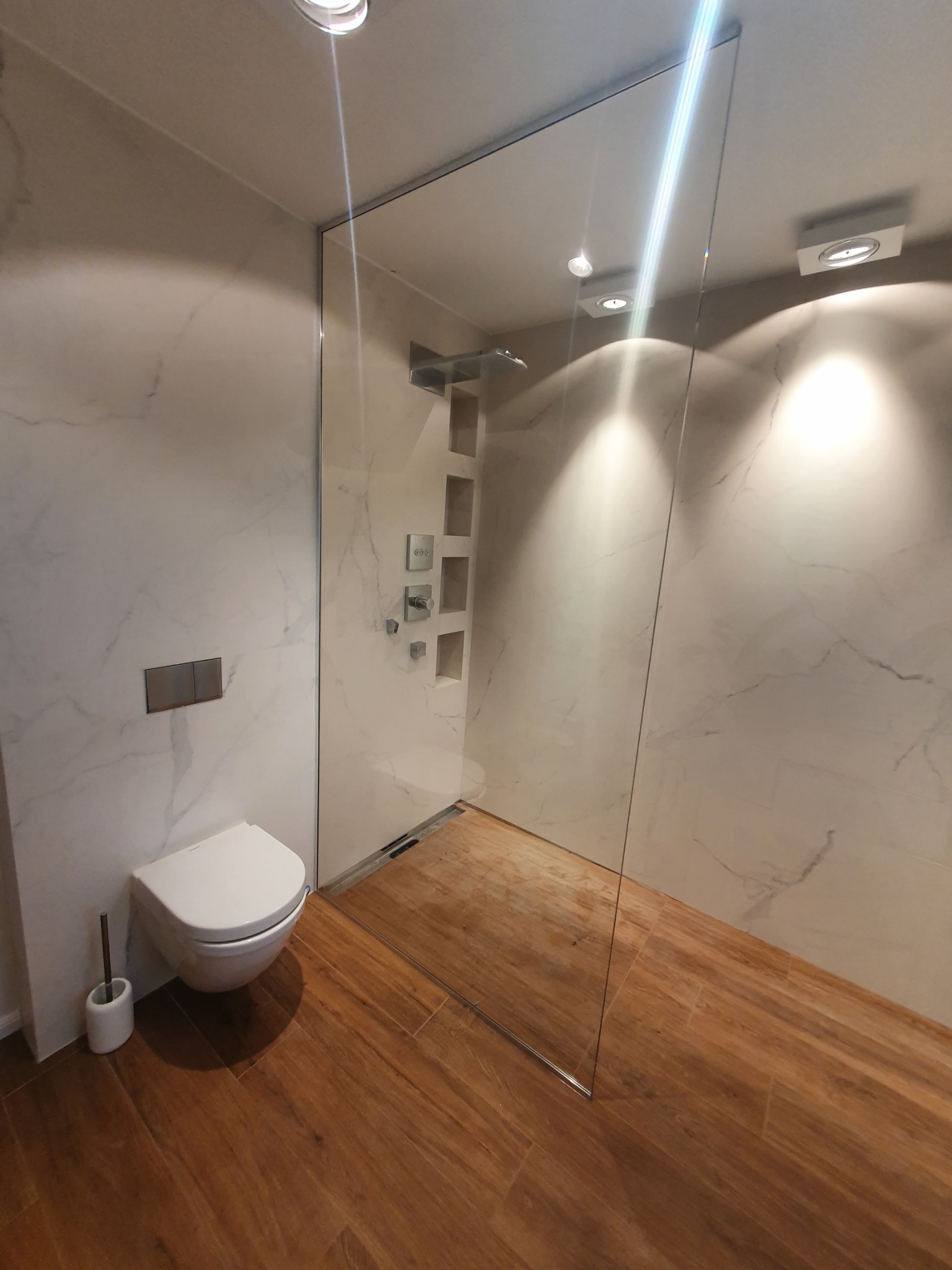 Nowoczesna łazienka z kabiną prysznicową typu walk-in, wykończona marmurowymi płytami i drewnopodobną podłogą, z widoczną armaturą i toaletą podwieszaną.