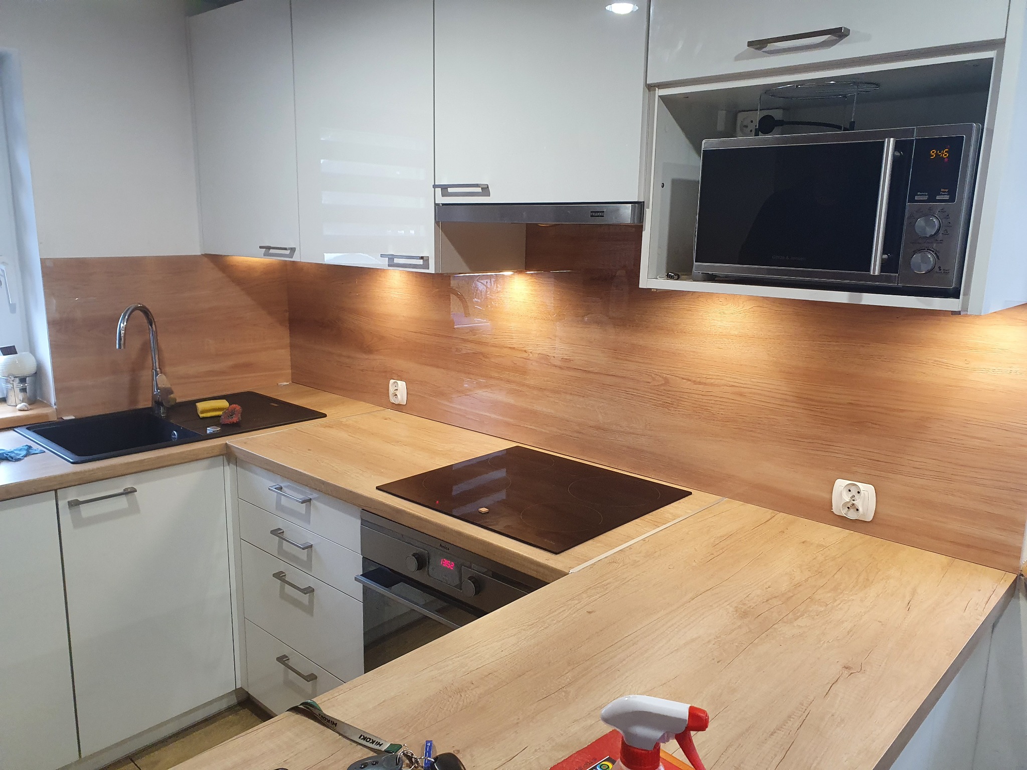 Nowoczesna kuchnia z białymi szafkami i blatem imitującym drewno, czarny zlew i płyta indukcyjna, mikrofala w zabudowie, widoczne gniazdka elektryczne na ścianie.