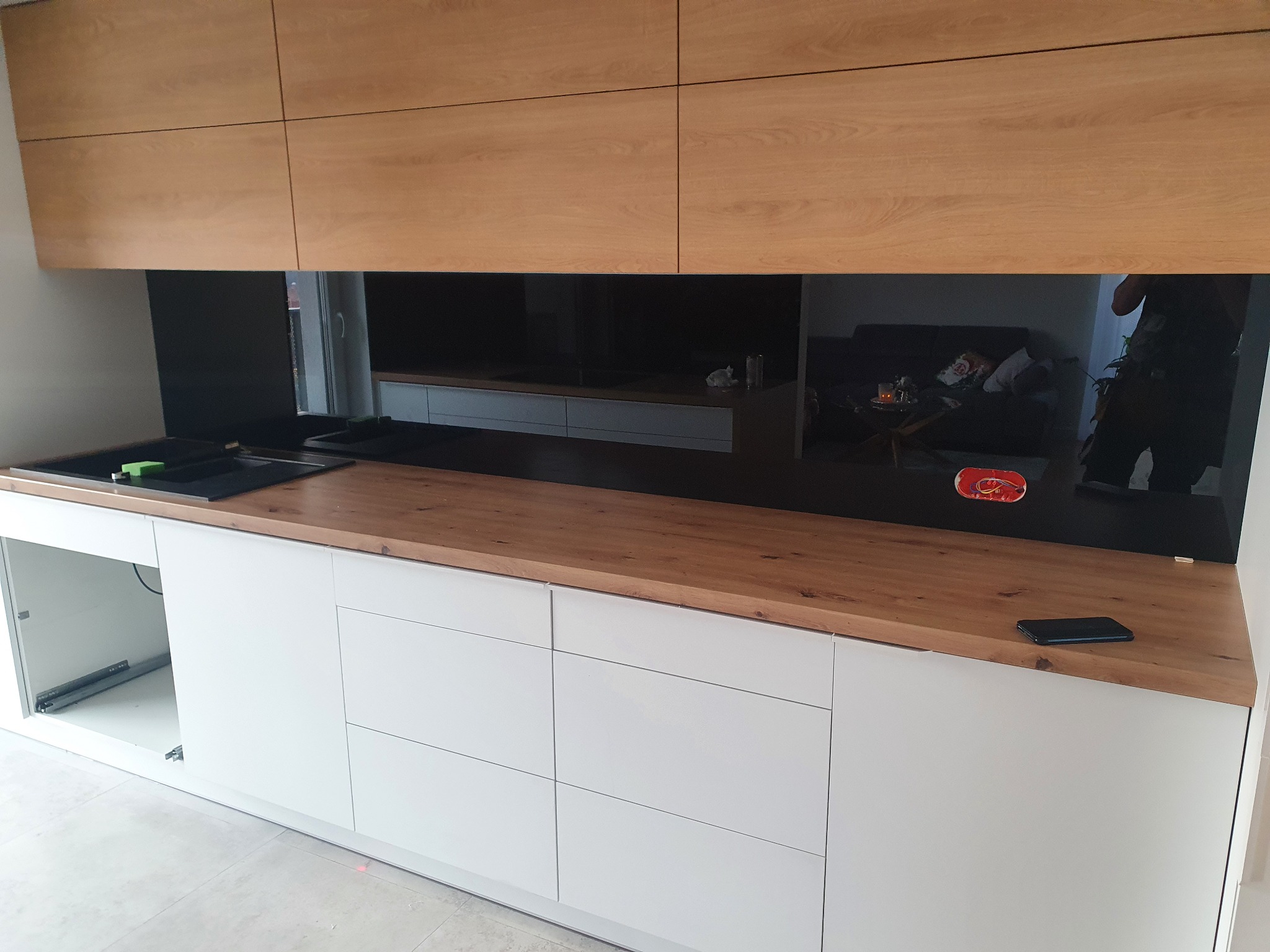 Nowoczesna kuchnia z białymi szafkami i drewnianym blatem, nad którym zamontowano czarny panel szklany na ścianie, odbijający wnętrze salonu.